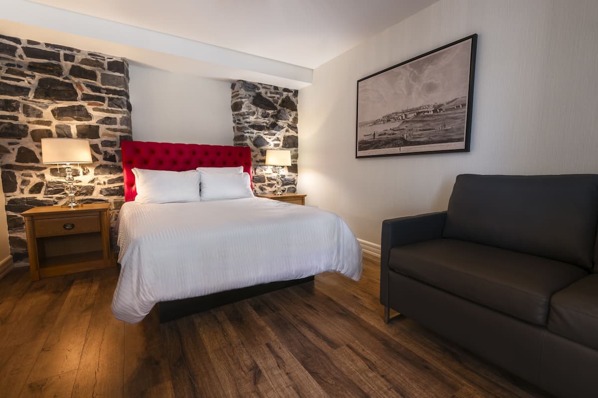 A bedroom with a stone wall, a red headboard, a bed with white bedding, two bedside lamps, a dark sofa, and a framed picture on the wall.