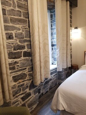 A room with a stone wall, a window with curtains, a bed, and a small wooden stool. - Auberge du Tresor (Québec City)