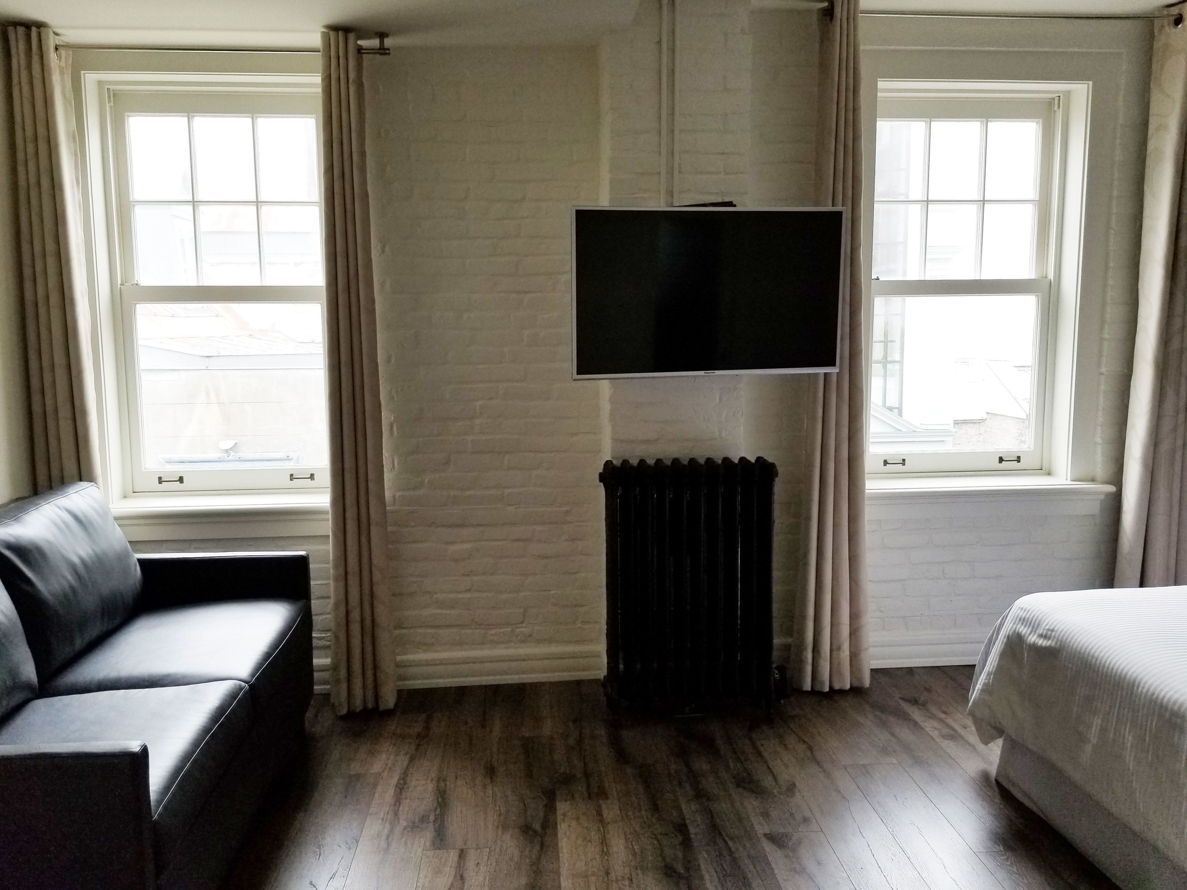 A hotel room with a black leather sofa, a bed, a flat-screen TV mounted on a brick wall, and a radiator.