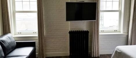 A hotel room with a black leather sofa, a bed, a flat-screen TV mounted on a brick wall, and a radiator.