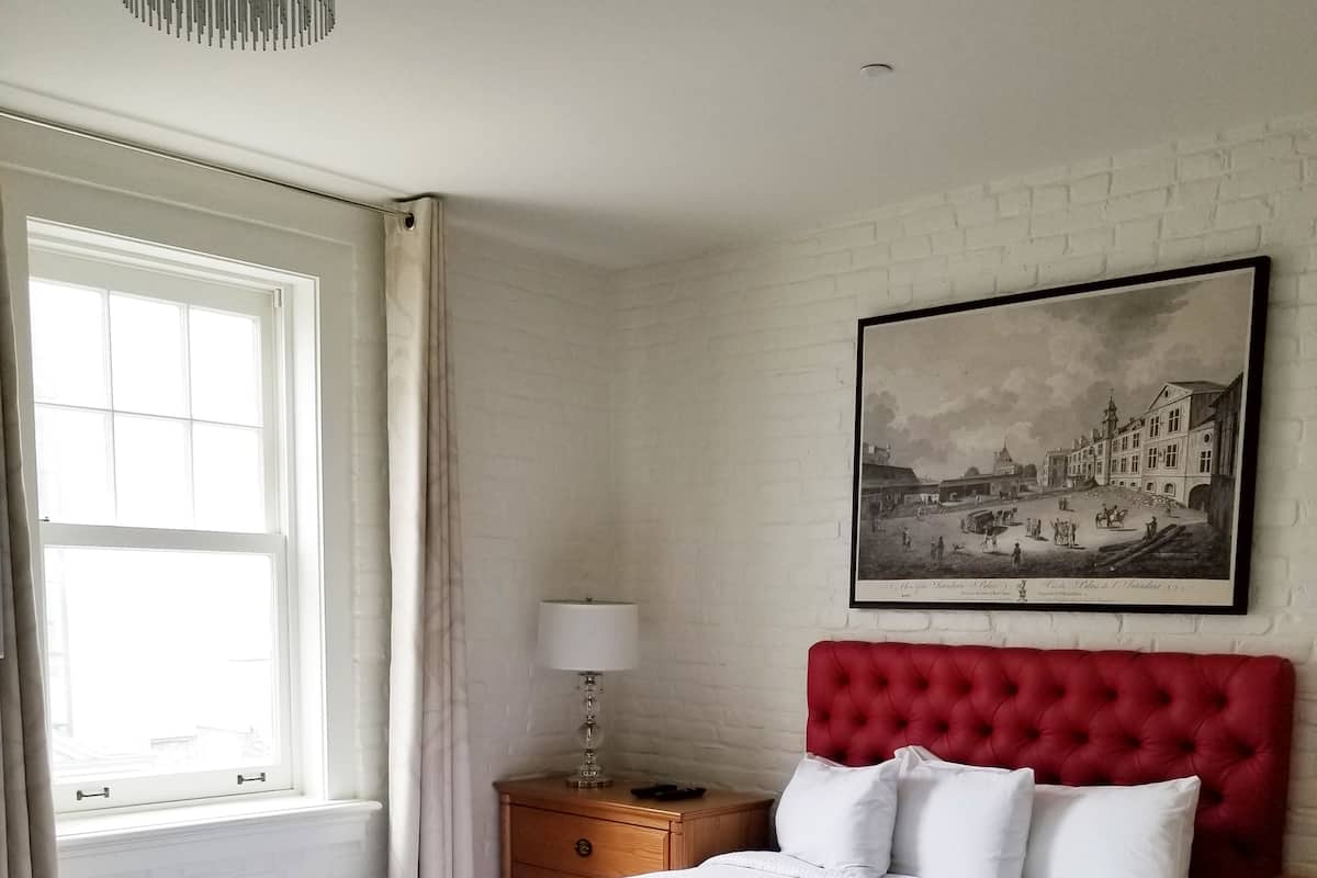 A neatly arranged bedroom with a large bed, a wooden nightstand, a red upholstered headboard, a framed picture on the wall, and a window with curtains.