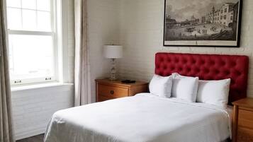 Un dormitorio ordenado con una cama grande, una mesita de noche de madera, un cabecero tapizado rojo, un cuadro enmarcado en la pared y una ventana con cortinas.