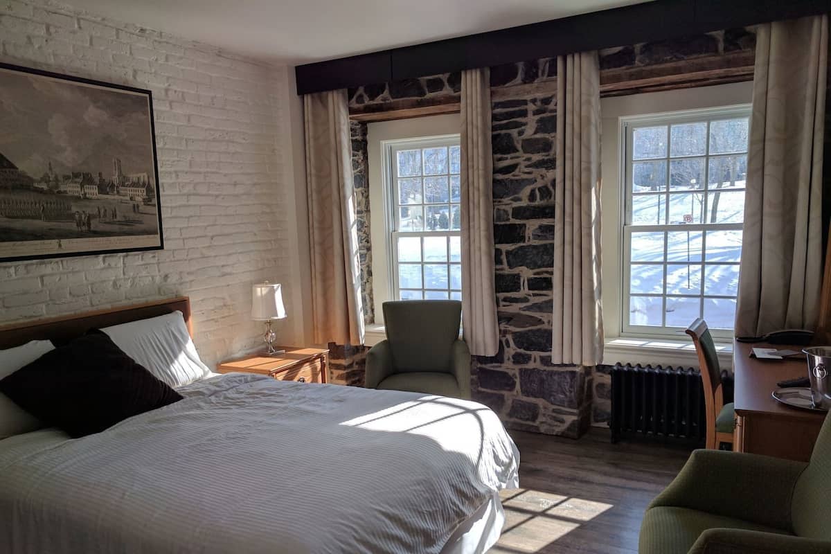 A hotel room with a large bed, a chair, a desk, a radiator, and a stone wall.