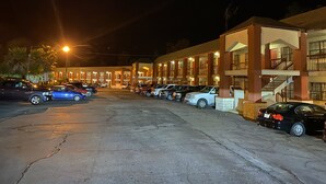 A multi-story hotel with a parking lot filled with cars. The building has multiple balconies and a staircase on the right side.