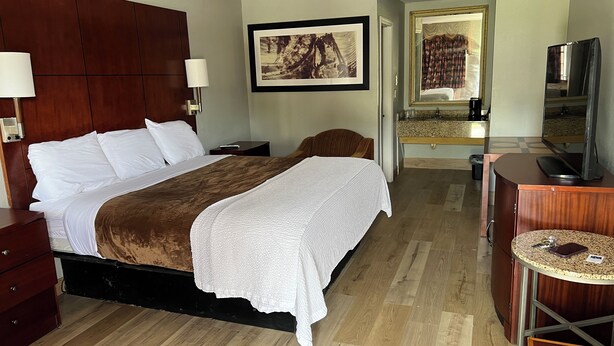 A hotel room with a large bed, wooden headboard, a bedside table, a TV, a small round table, and a mirror.