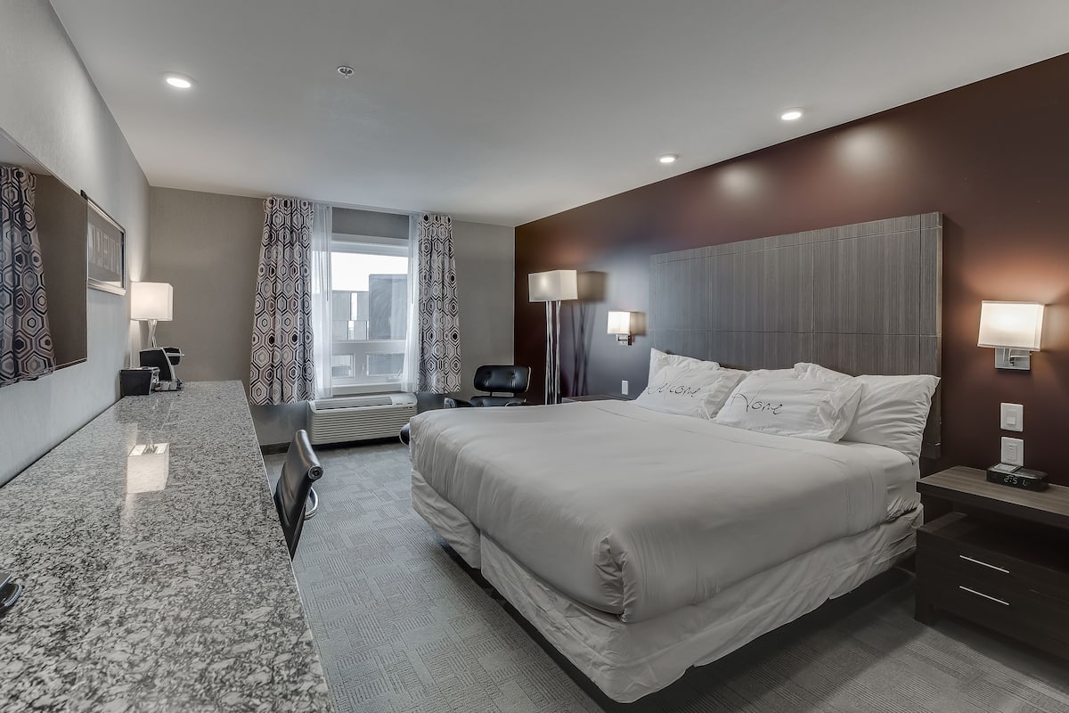 Une chambre d’hôtel moderne, dotée d’un grand lit, d’un bureau et d’une chaise. Une fenêtre, ornée de rideaux, offre une vue sur la ville.