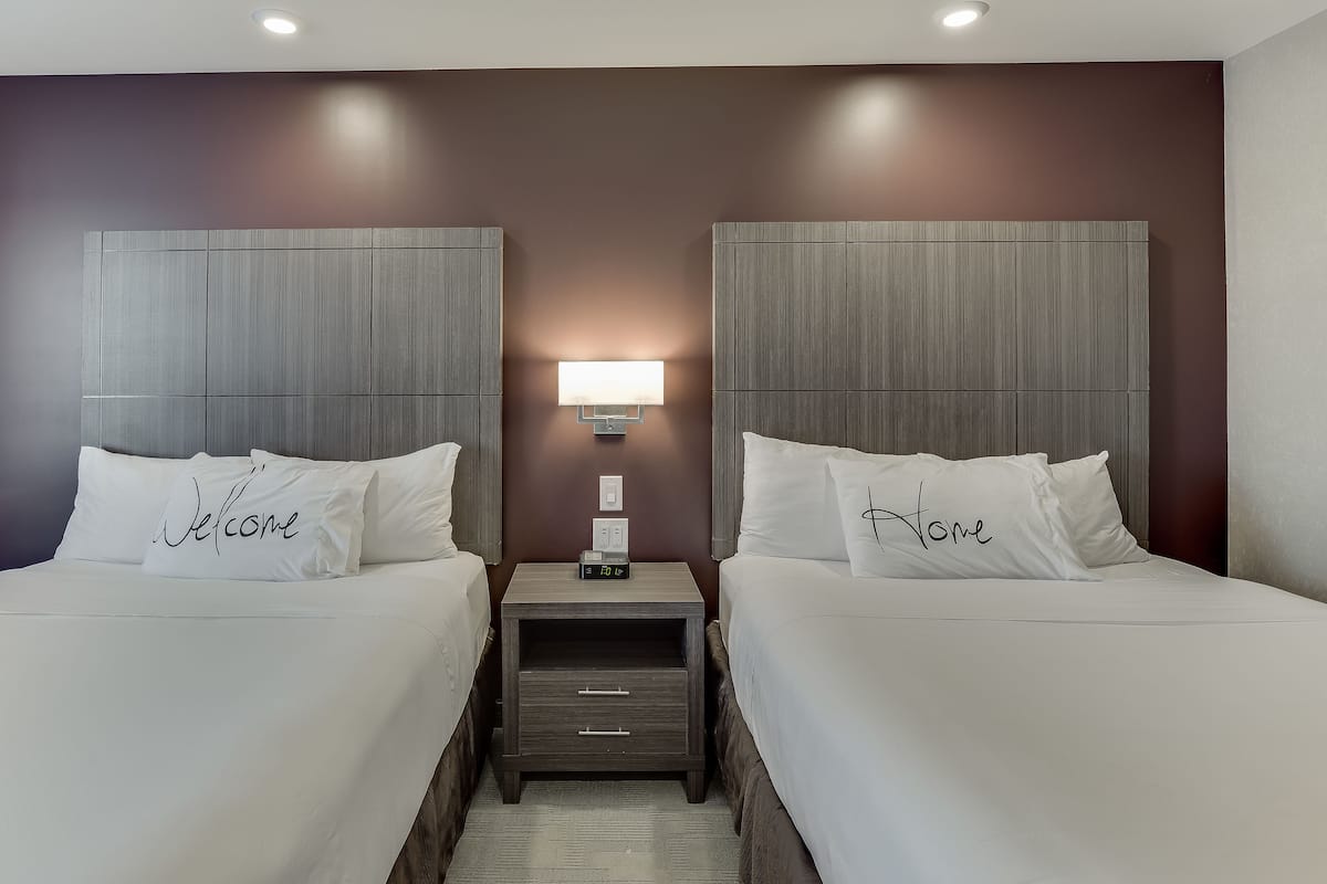 Une chambre d’hôtel avec deux lits, chacun agrémenté d’un oreiller portant l’inscription « Bienvenue » et « Chez vous ». Une table de chevet avec une lampe et une horloge numérique se trouve entre les deux lits.