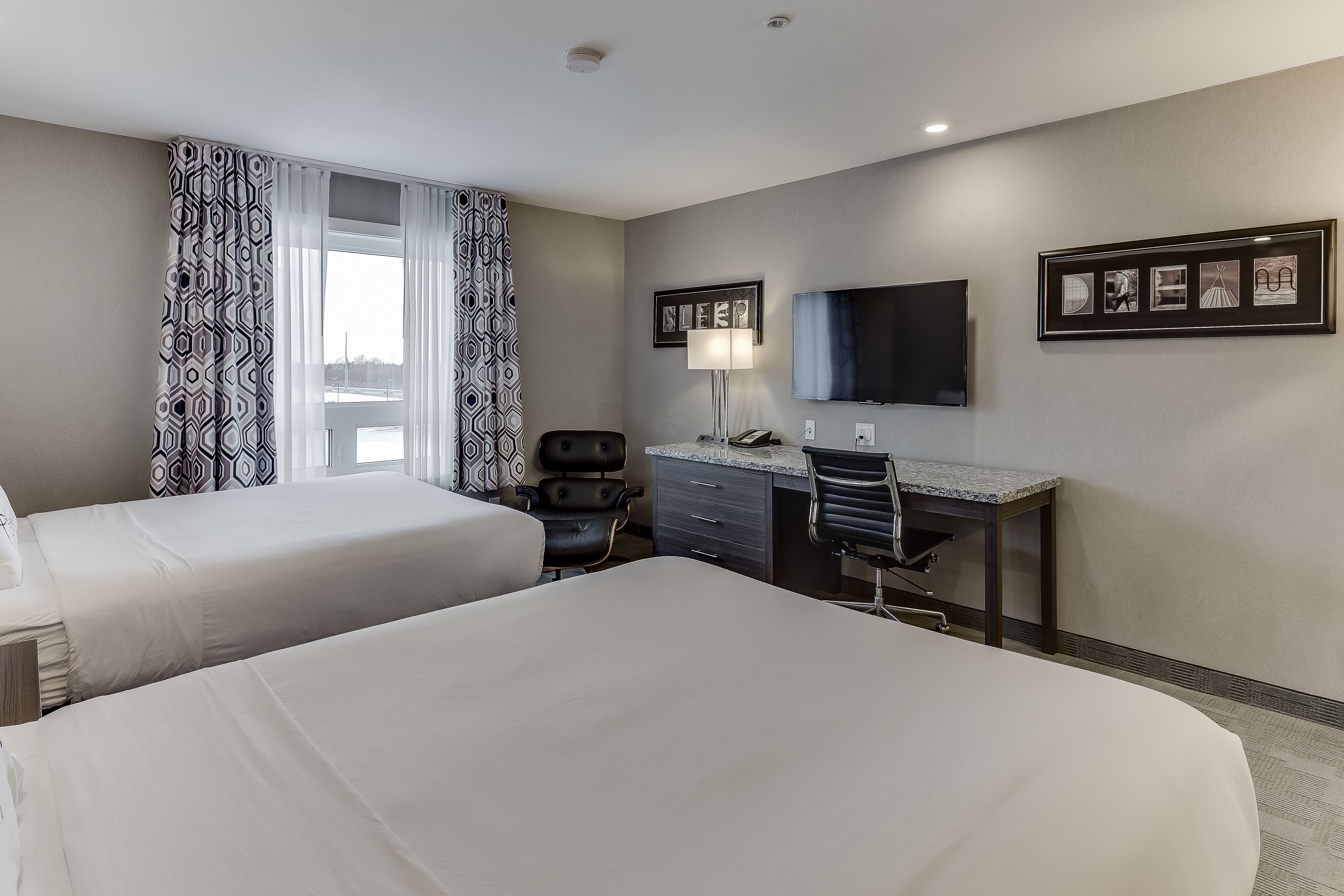 A hotel room with two beds, a desk, a chair, a TV, and a window with patterned curtains.