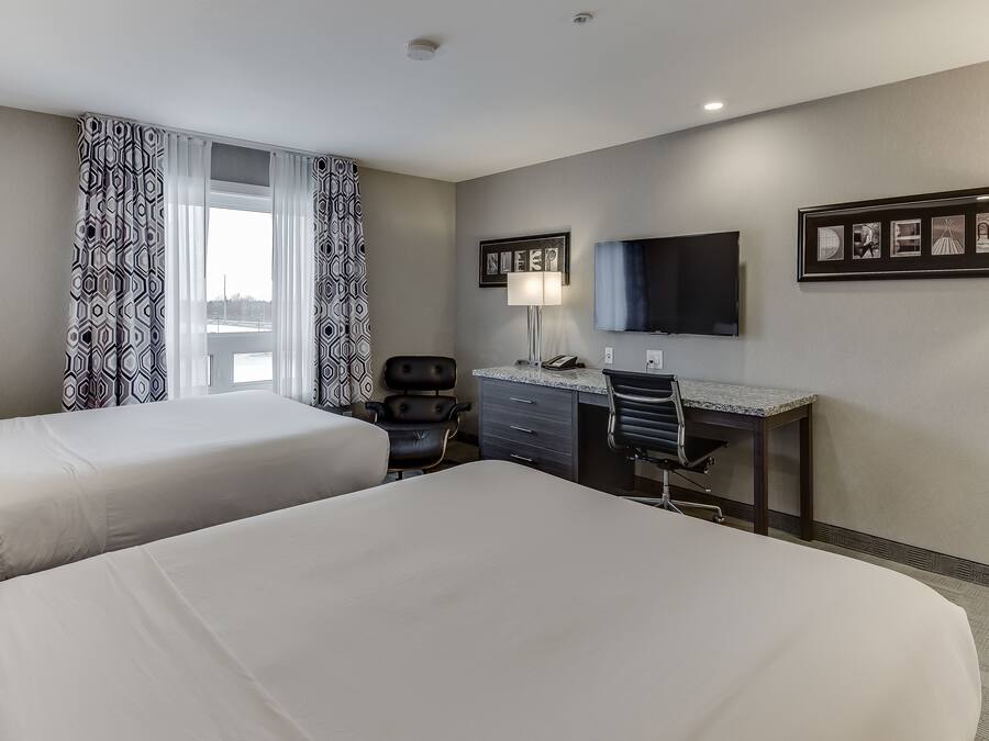 A hotel room with two beds, a desk, a chair, a TV, and a window with patterned curtains.