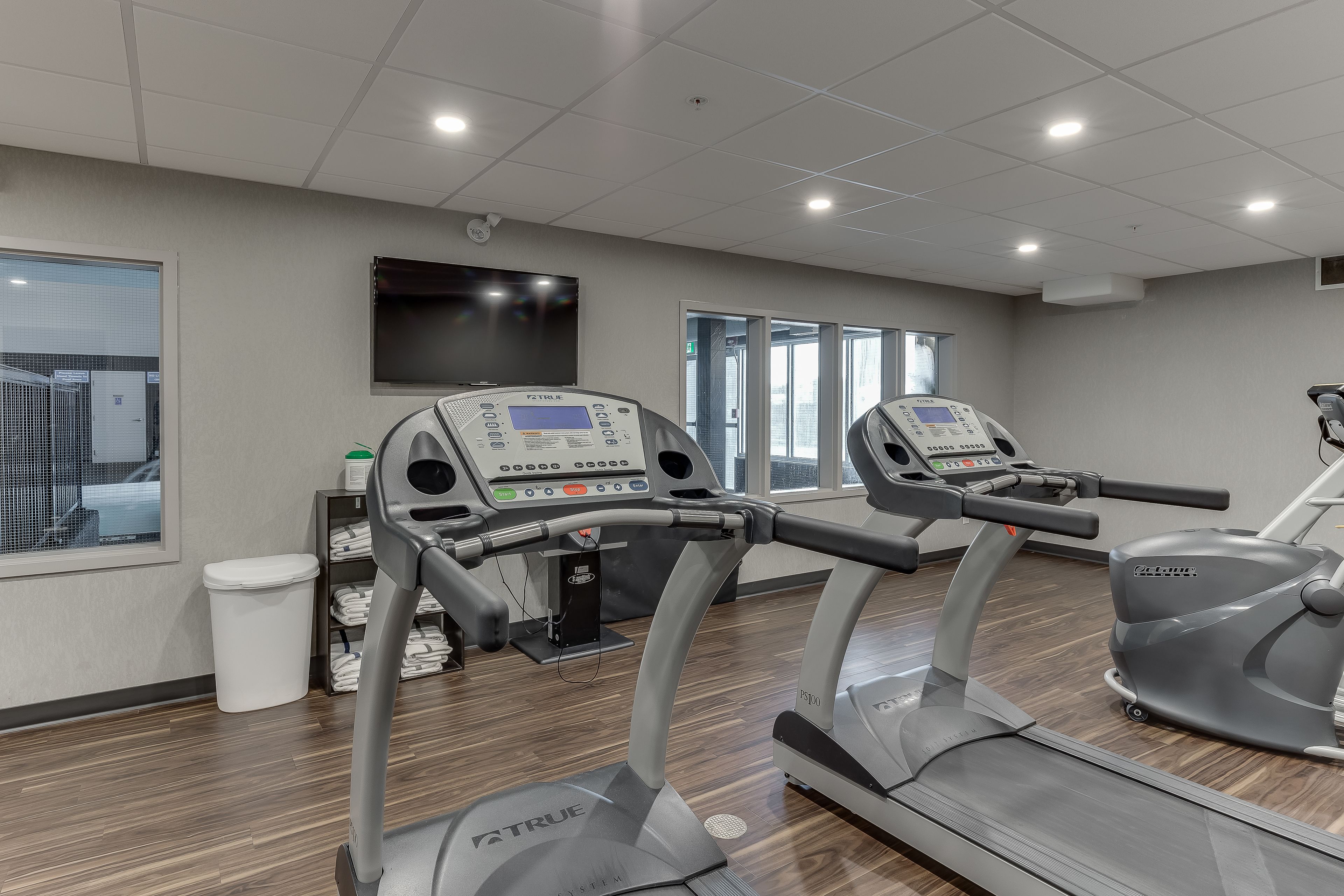 A gym with treadmills and an elliptical machine, a TV on the wall, and a window with a view of buildings.