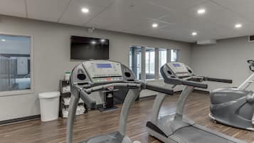 A gym with treadmills and an elliptical machine, a TV on the wall, and a window with a view of buildings.