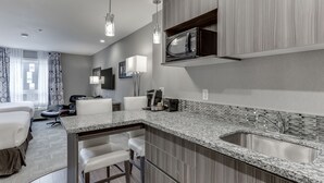Cocina moderna con encimera de granito, pileta de acero inoxidable y microondas. Cuenta con un desayunador con taburetes y una sala de estar con sofá y sillones.
