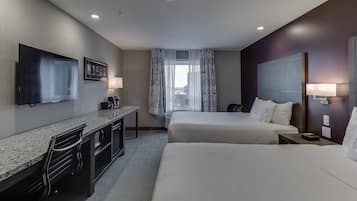 Ein Hotelzimmer mit einem großen Bett, einem kleineren Bett, einem Schreibtisch mit Stuhl, einem Flachbildfernseher und einem Fenster mit gemusterten Vorhängen.