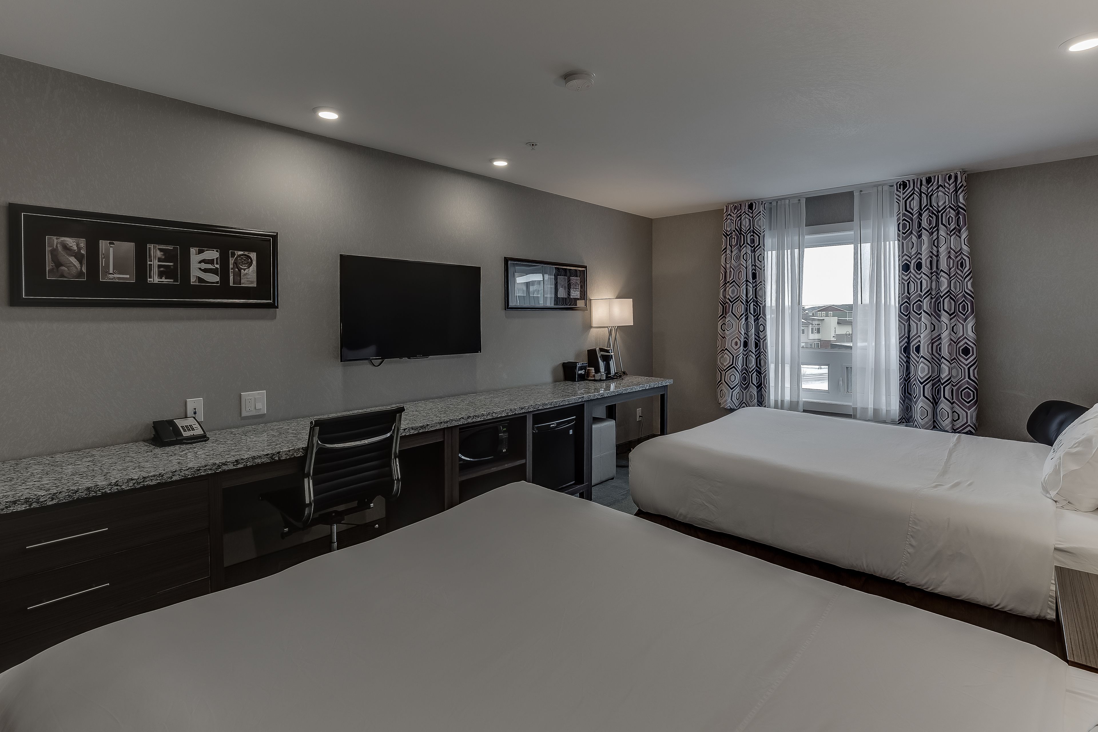 A hotel room with two beds, a desk with a flat-screen TV, a lamp, and a window with patterned curtains.