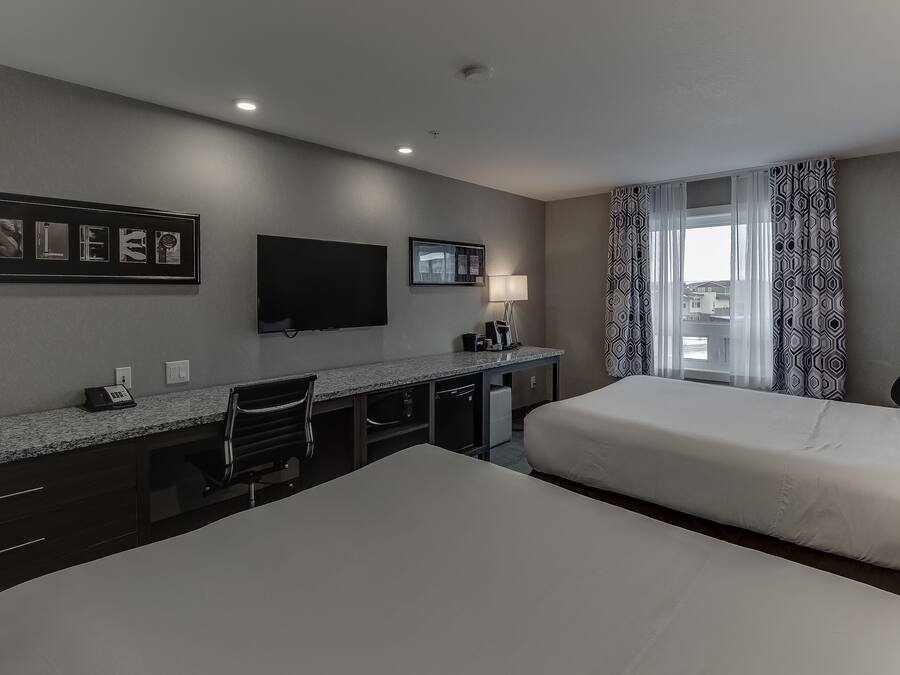 Ein Hotelzimmer mit zwei Betten, einem Schreibtisch, einem Flachbildfernseher, einer Lampe und einem Fenster mit gemusterten Vorhängen.