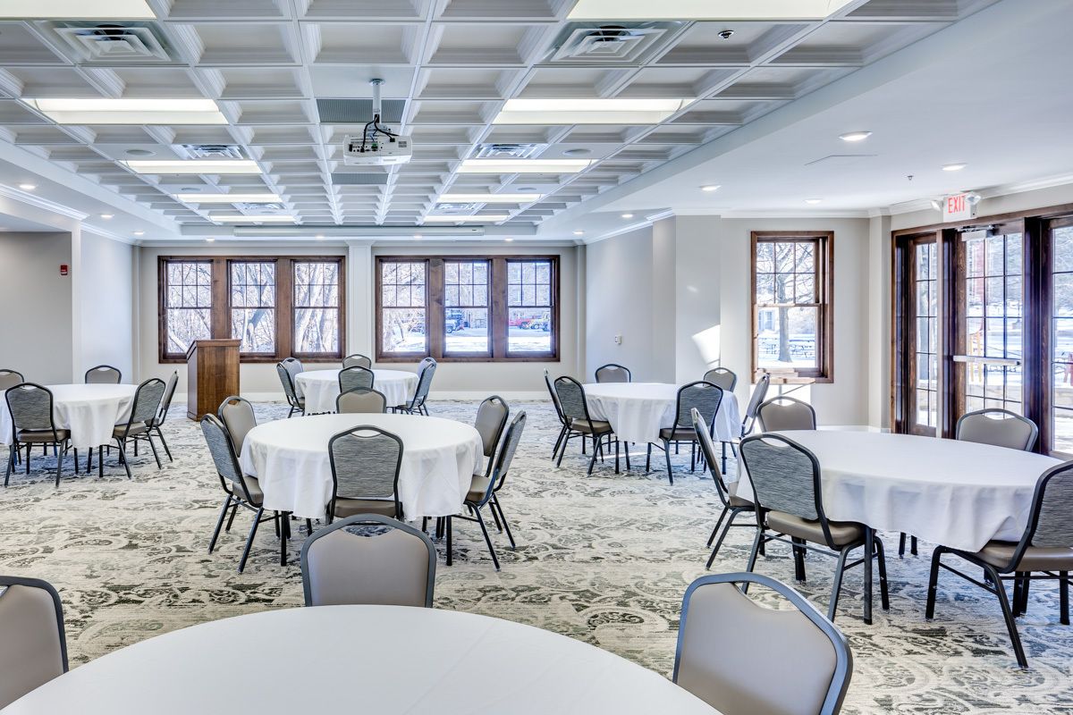 A conference room with multiple round tables and chairs, large windows with views of the outside, and a patterned carpet.