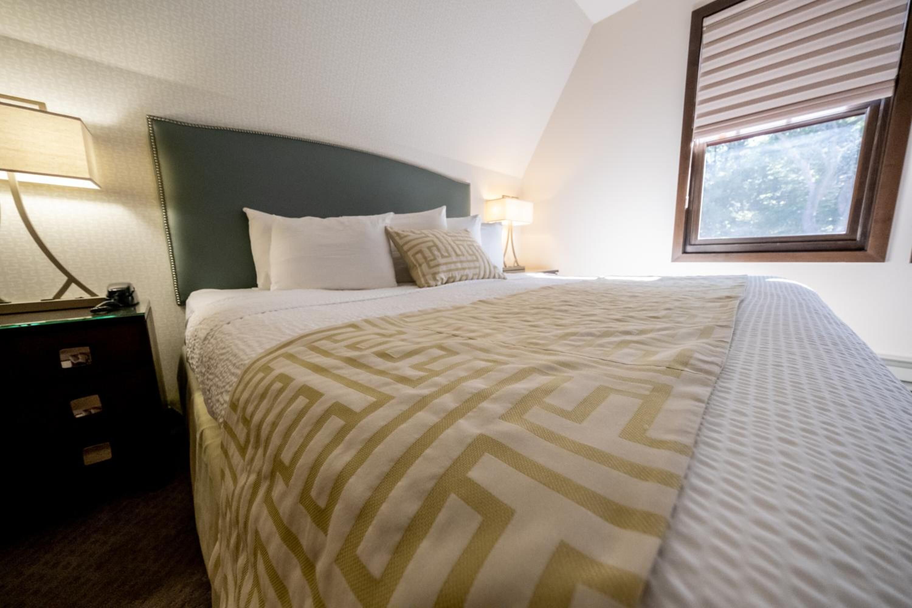 A neatly made bed with a patterned bedspread, a bedside lamp, and a window with blinds.