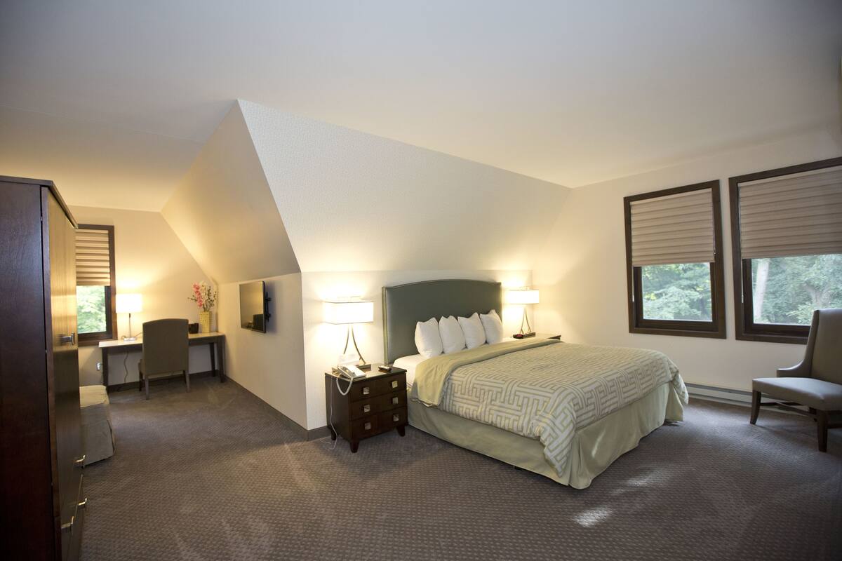 Habitación de hotel con cama, escritorio, silla y dos ventanas con persianas.