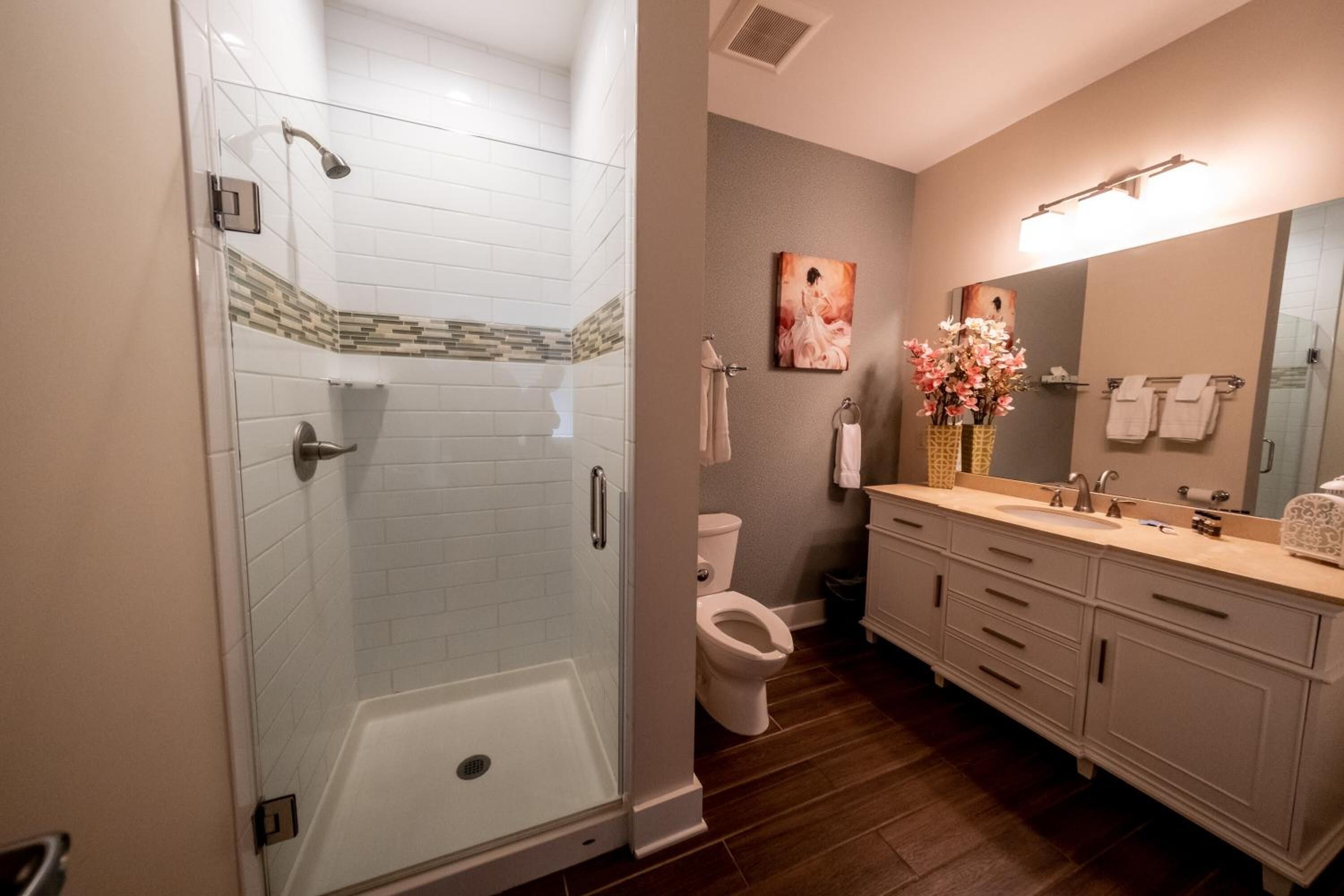 A bathroom with a glass-enclosed shower, a toilet, a sink with a mirror, and a vase of flowers.