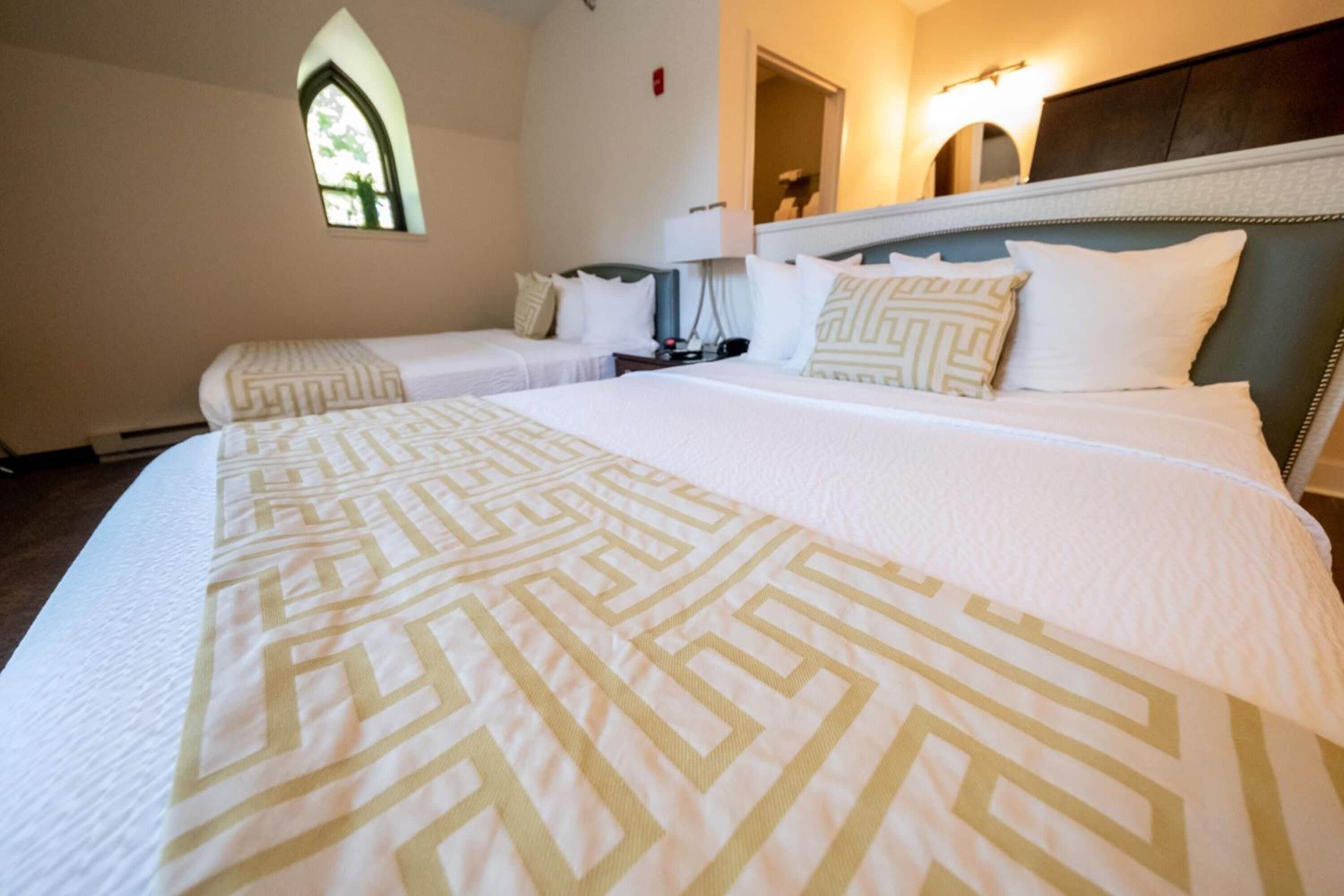 Habitación de hotel con dos camas individuales, una cama grande con cubrecama estampado, una mesita de noche, una lámpara y una ventana con vistas.