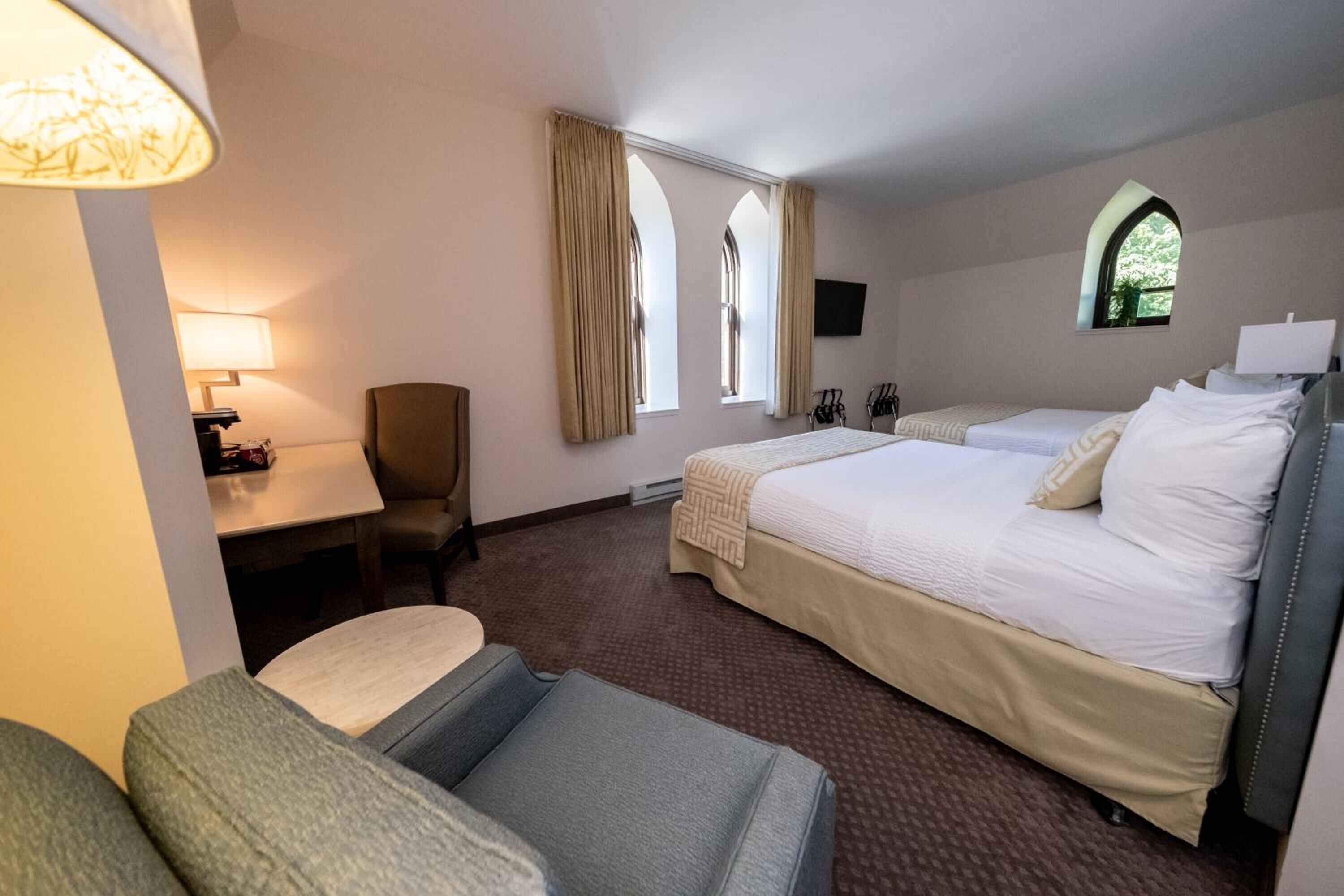 A hotel room with a bed, a desk, a chair, and two windows with curtains.