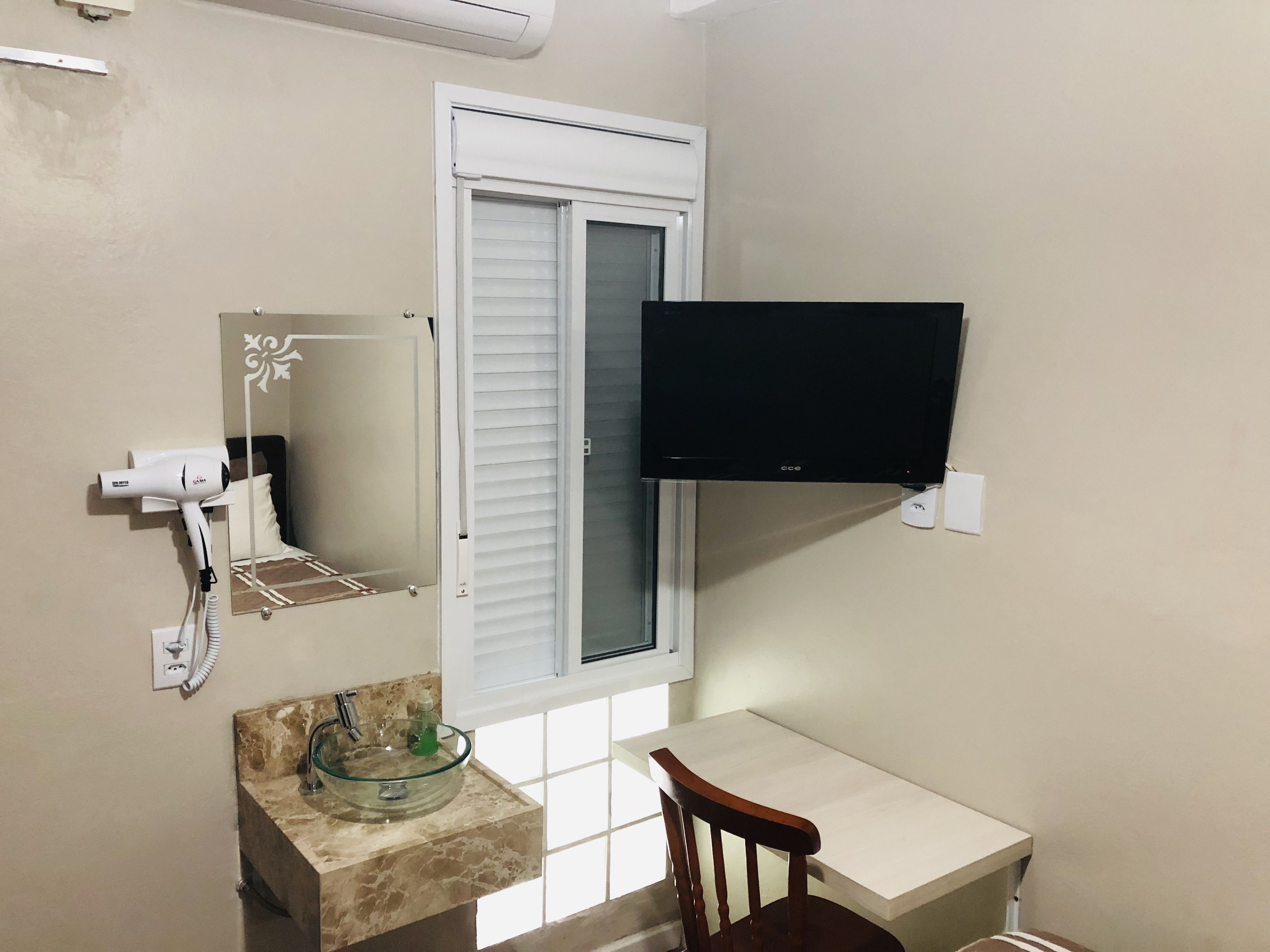 Un baño con lavamanos de vidrio, un secador de pelo, un espejo, una ventana con persianas y un televisor de pantalla plana montado en la pared.