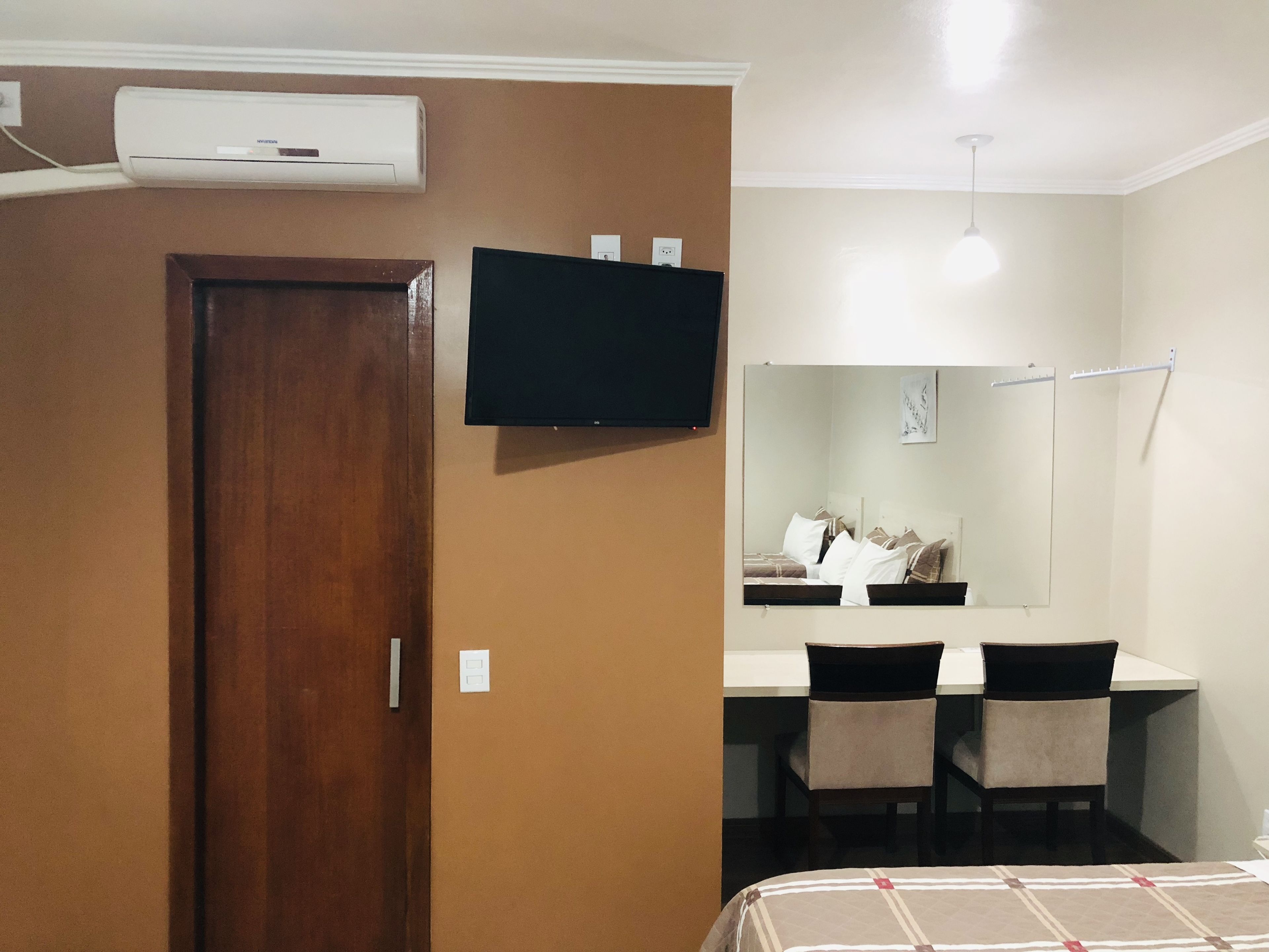 Una habitación con puerta de madera, televisor de pantalla plana, cama con cubrecama estampado, escritorio con dos sillas y un espejo en la pared.
