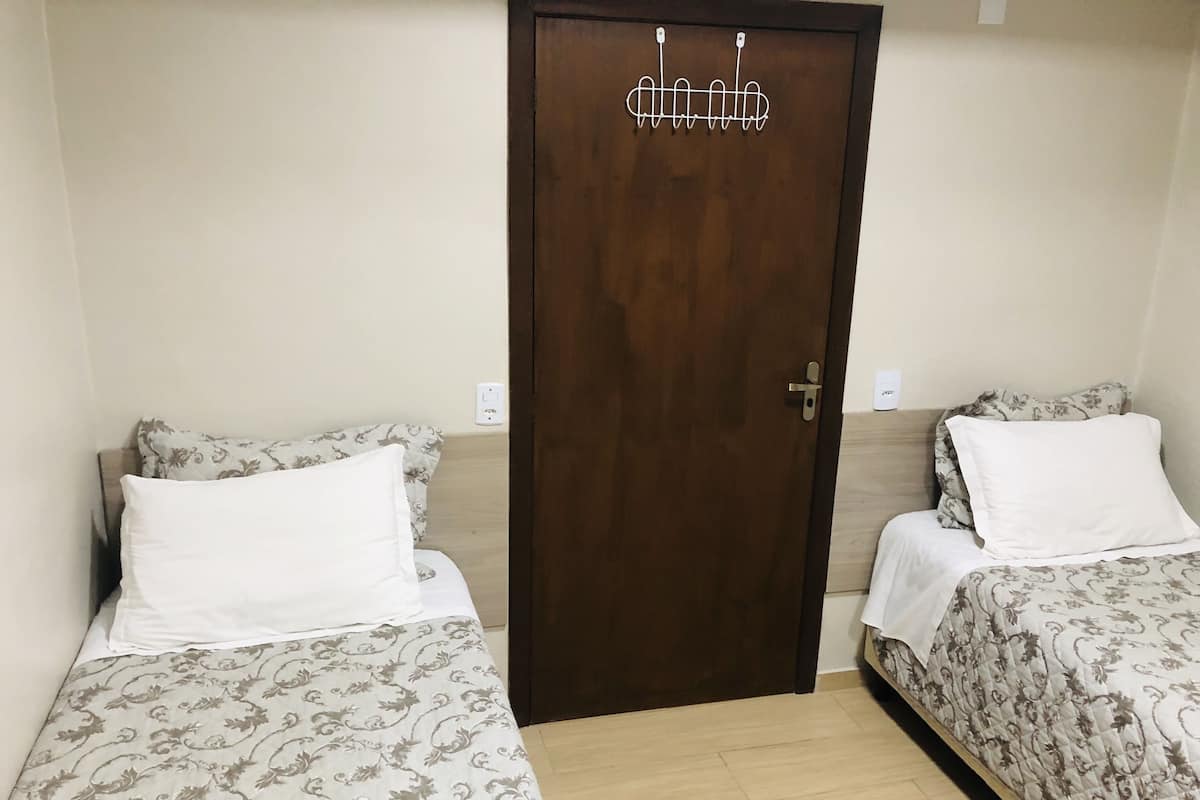 Ein Zimmer mit zwei Einzelbetten, jeweils mit gemusterten Bettbezügen und weißen Kissen, einer Holztür mit Kleiderhaken und einem hellen Boden.