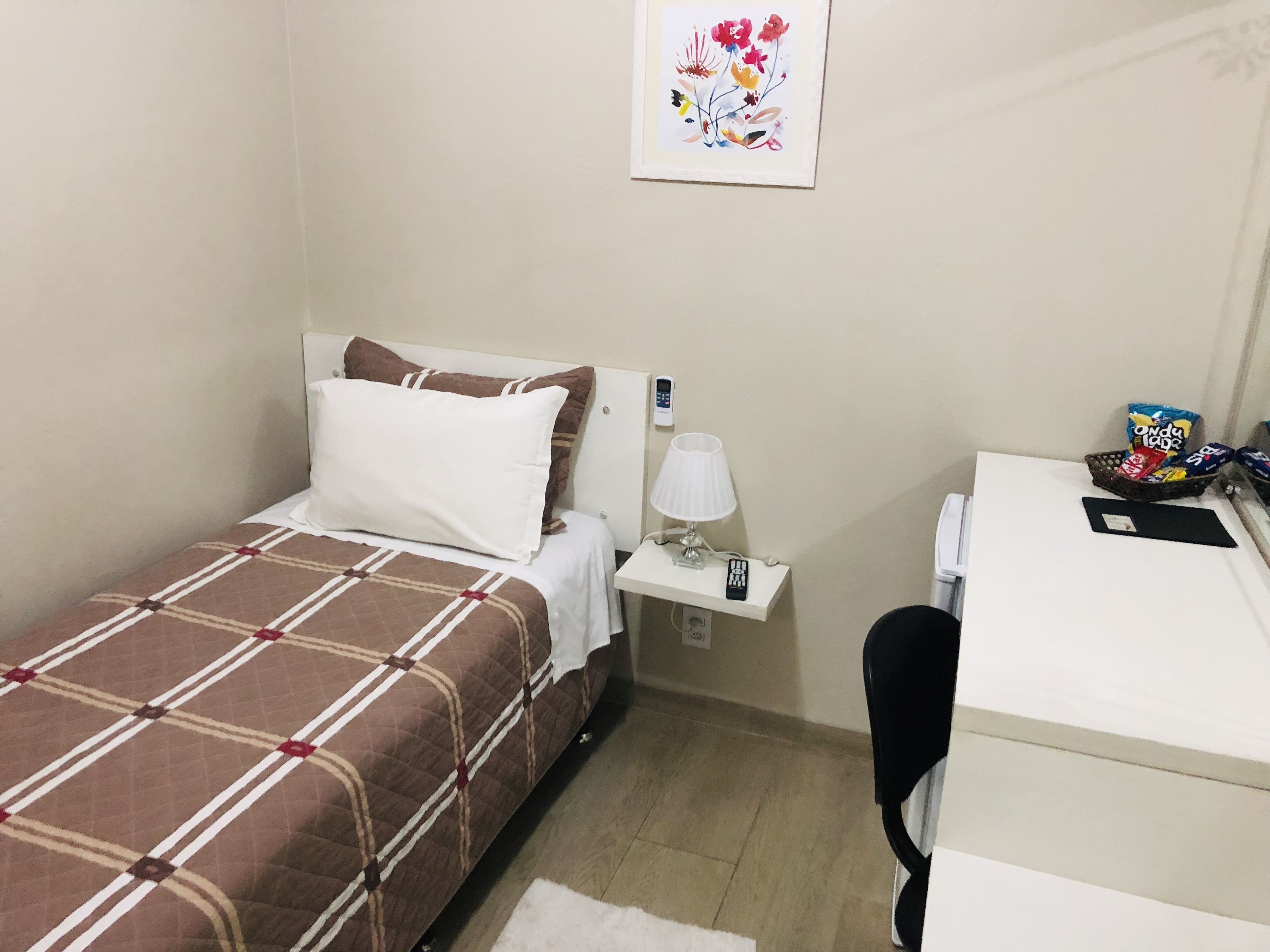 A small hotel room with a bed, a desk, a chair, a lamp, a phone, and a picture on the wall.