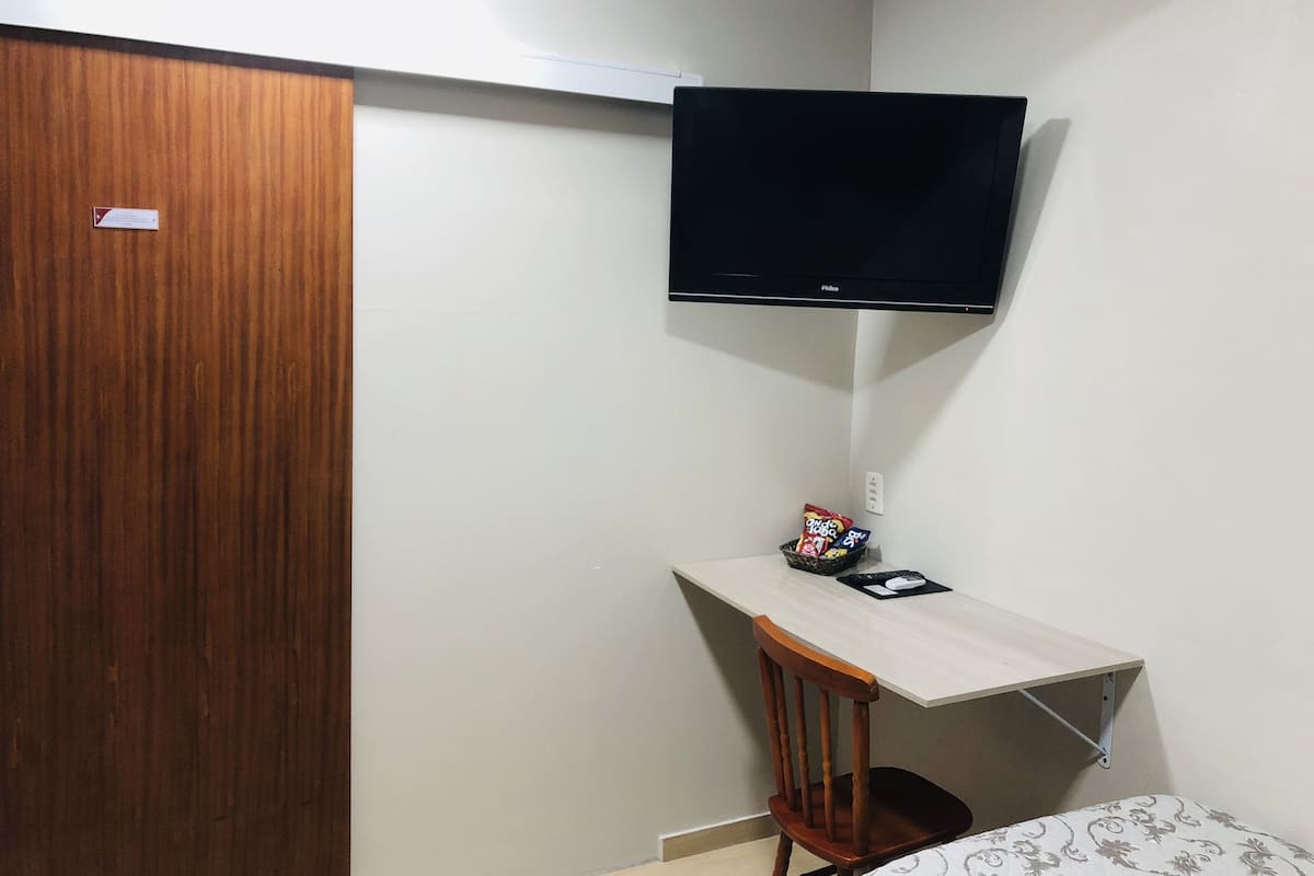 Ein kleines Zimmer mit einem Holzschrank, einem Schreibtisch mit Stuhl, einem an der Wand befestigten Fernseher und einem Bett mit gemusterten Betttextilien.