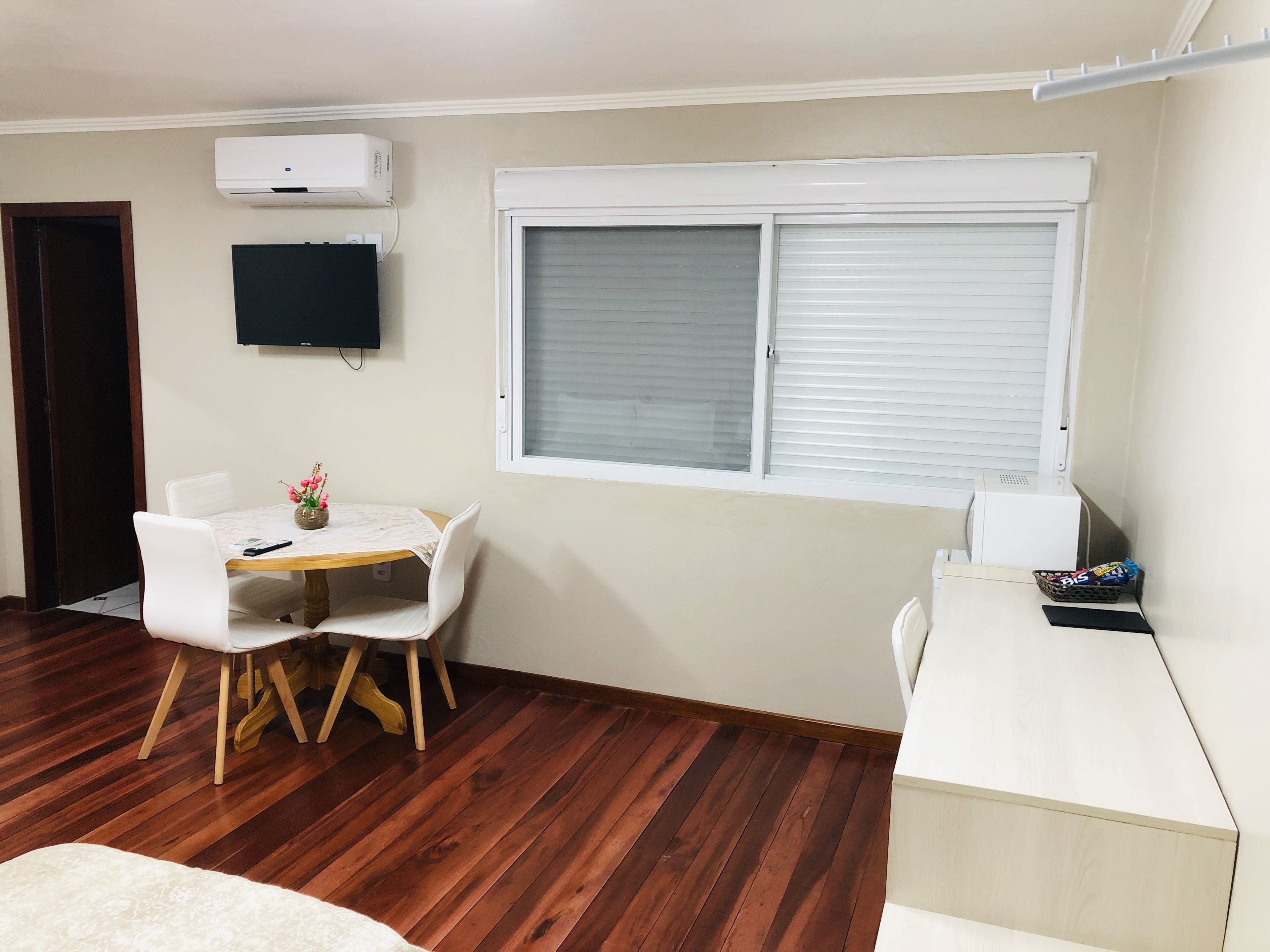 Ein Zimmer mit Holzboden, einem Esstisch mit Stühlen, einem an der Wand befestigten Fernseher und einem Fenster mit Jalousien.