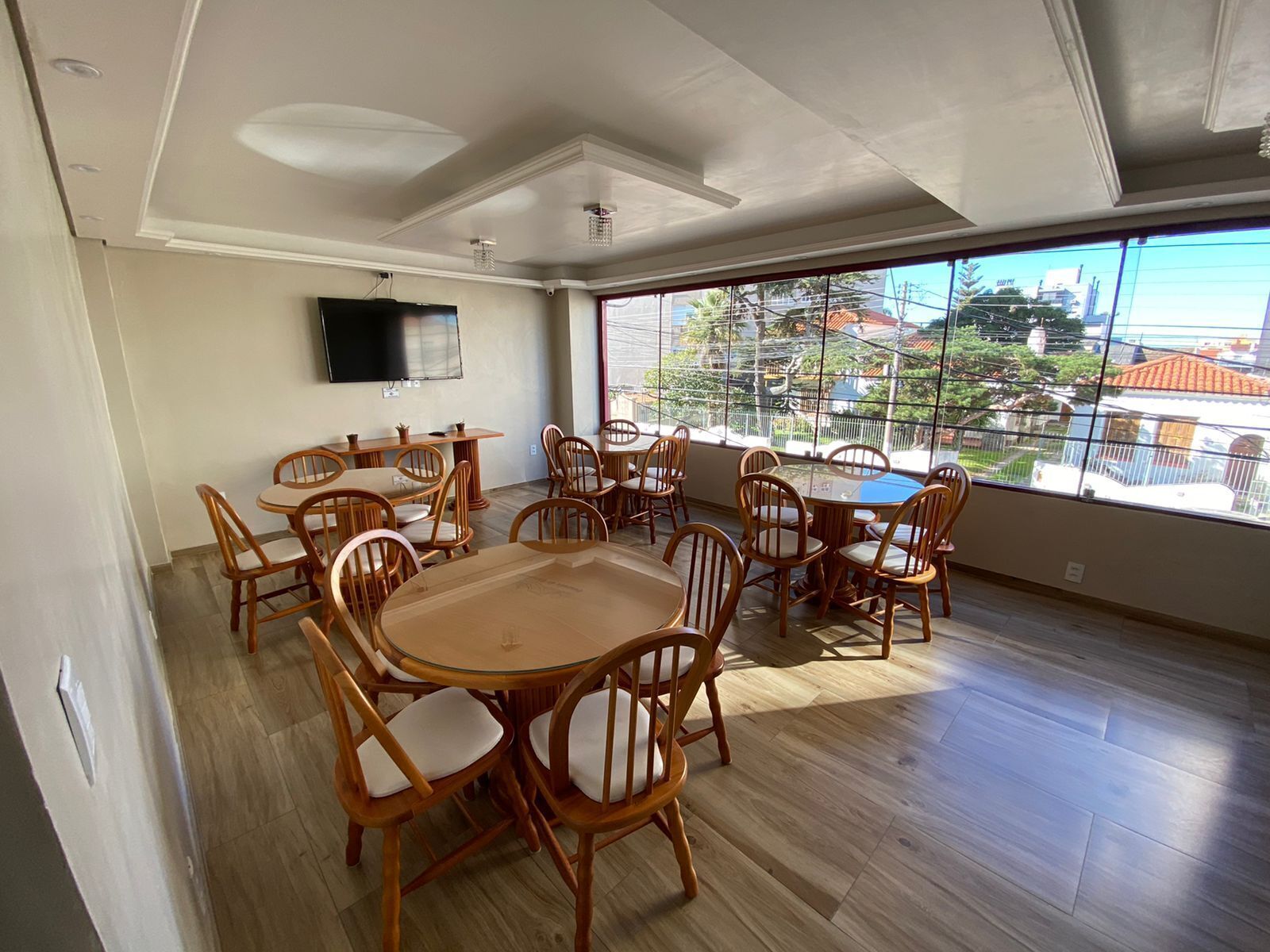 A spacious dining area with wooden tables and chairs, a flat-screen TV mounted on the wall, and large windows offering a view of the cityscape.