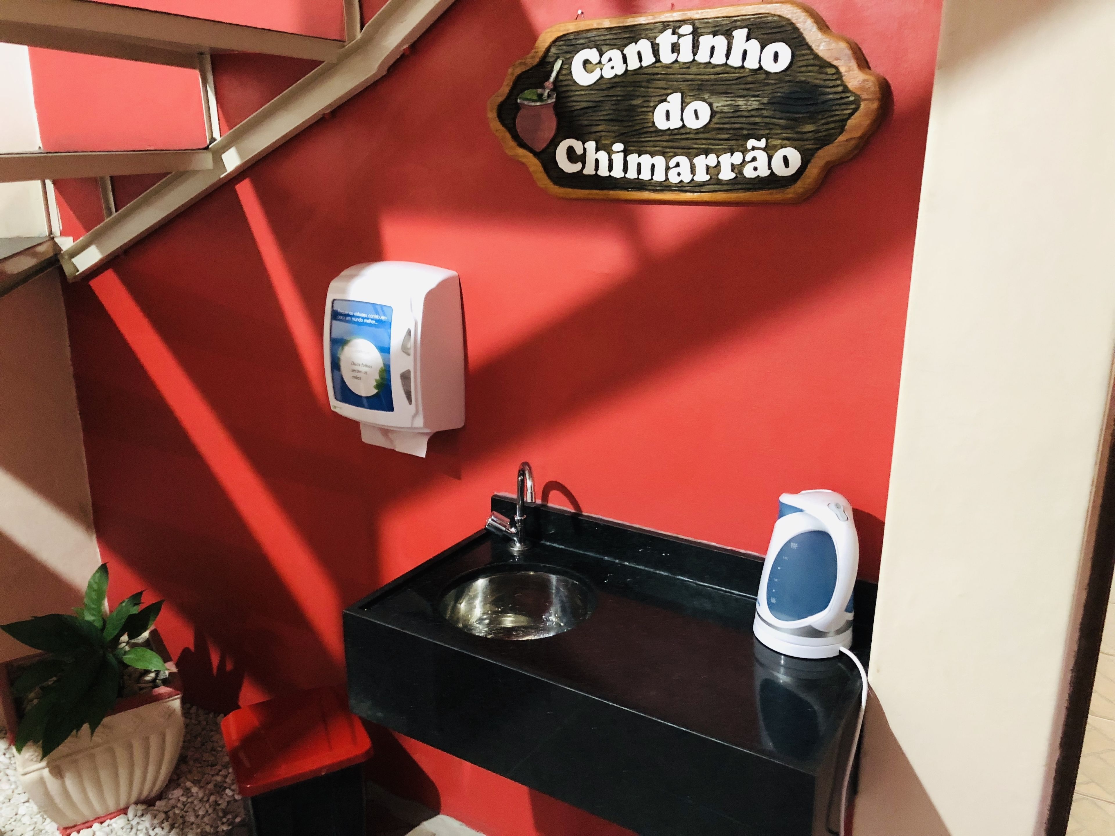 Un bagno con una parete rossa, un cartello di legno con su scritto "Cantinho do Chimarrão", un asciugatore per le mani, un lavandino e un bollitore.