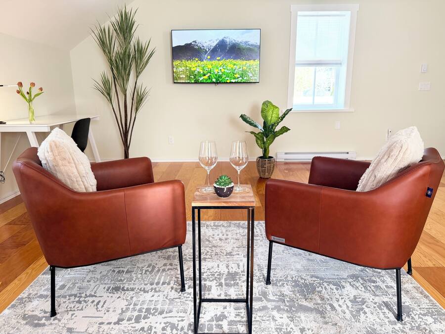 A living room with two brown leather chairs, a small table with two wine glasses, a potted plant, and a TV on the wall.