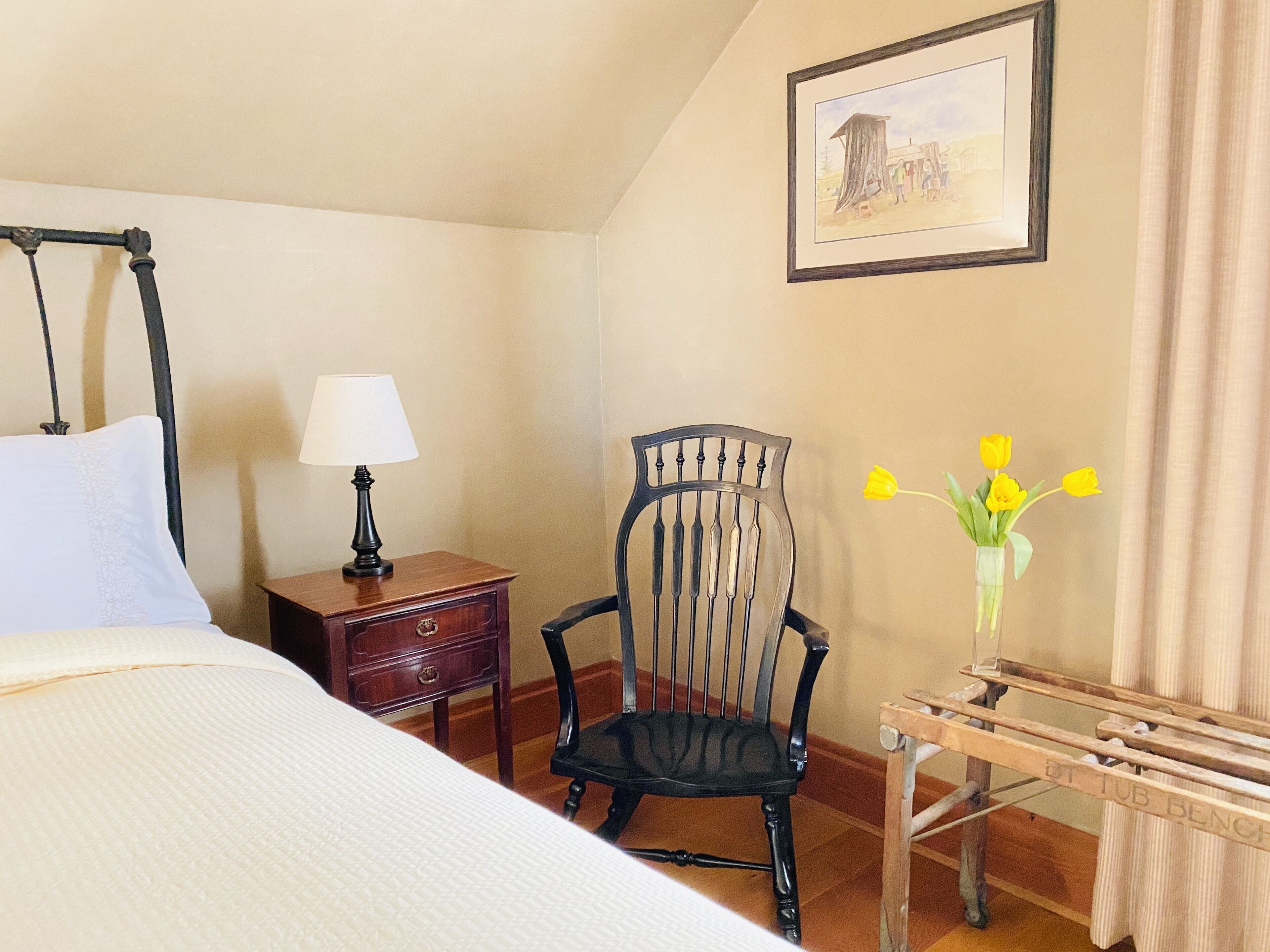 A bedroom with a four-poster bed, a wooden nightstand with a lamp, a chair, and a small table with a vase of flowers.