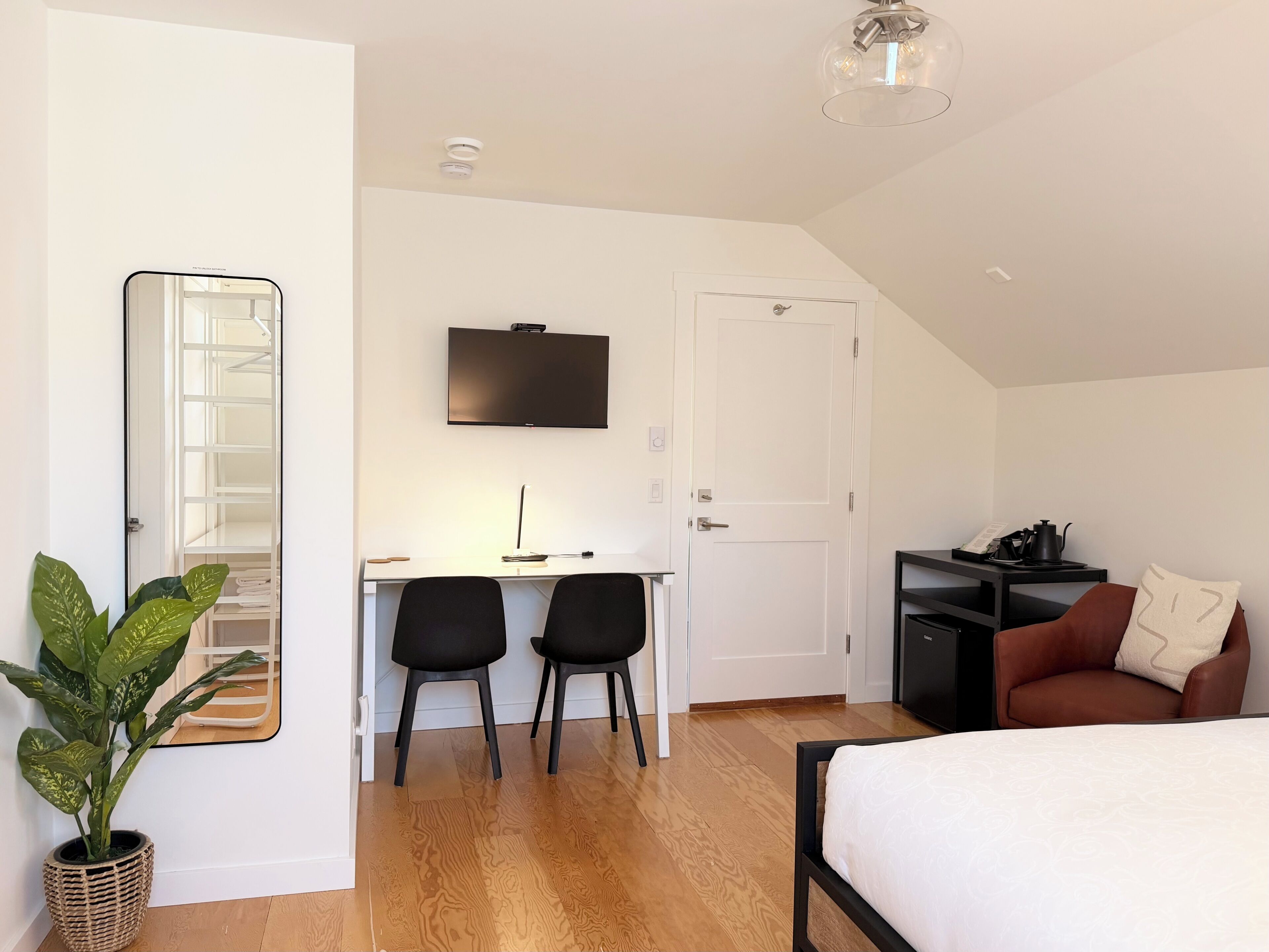 A modern bedroom with a bed, a desk with two chairs, a TV, and a potted plant.