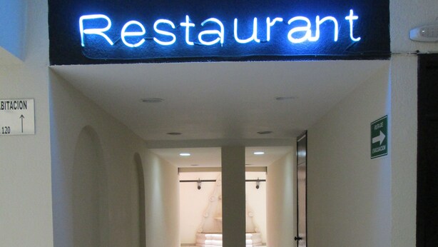 A hallway leading to a restaurant with a neon sign, a directional sign, and an emergency exit sign. - Hotel San Francisco (Culiacan)