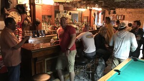Zona de bar con mesa de billar, taburetes y varios botellas y vasos en estantes.