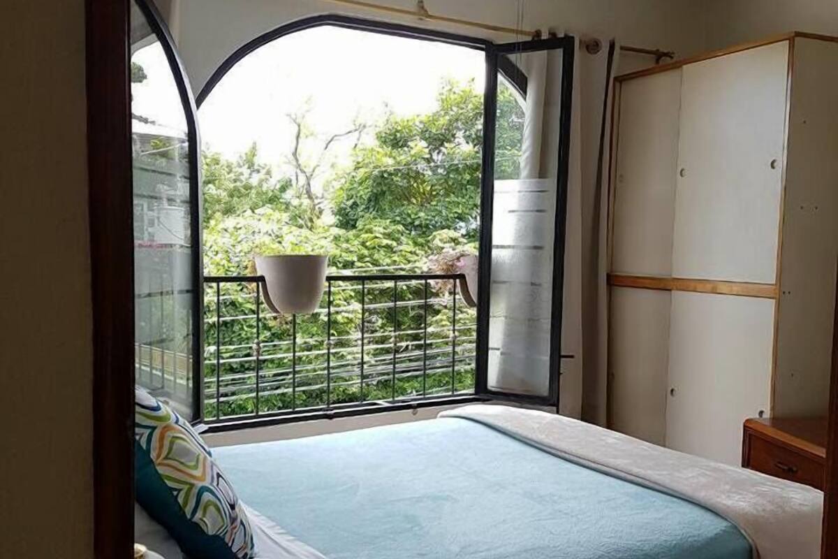 A bedroom with a bed, a bedside table, a wardrobe, and a balcony with a view of greenery.