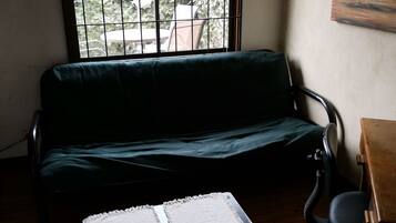 Une pièce avec un canapé vert, une petite table et une chaise. Il y a une fenêtre avec des barreaux en métal, et de là, on peut voir la verdure à l’extérieur.