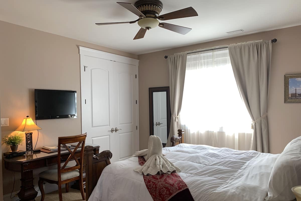 Une chambre d’hôtel avec un lit, un bureau avec une lampe, une chaise, un ventilateur de plafond et une fenêtre avec des rideaux.
