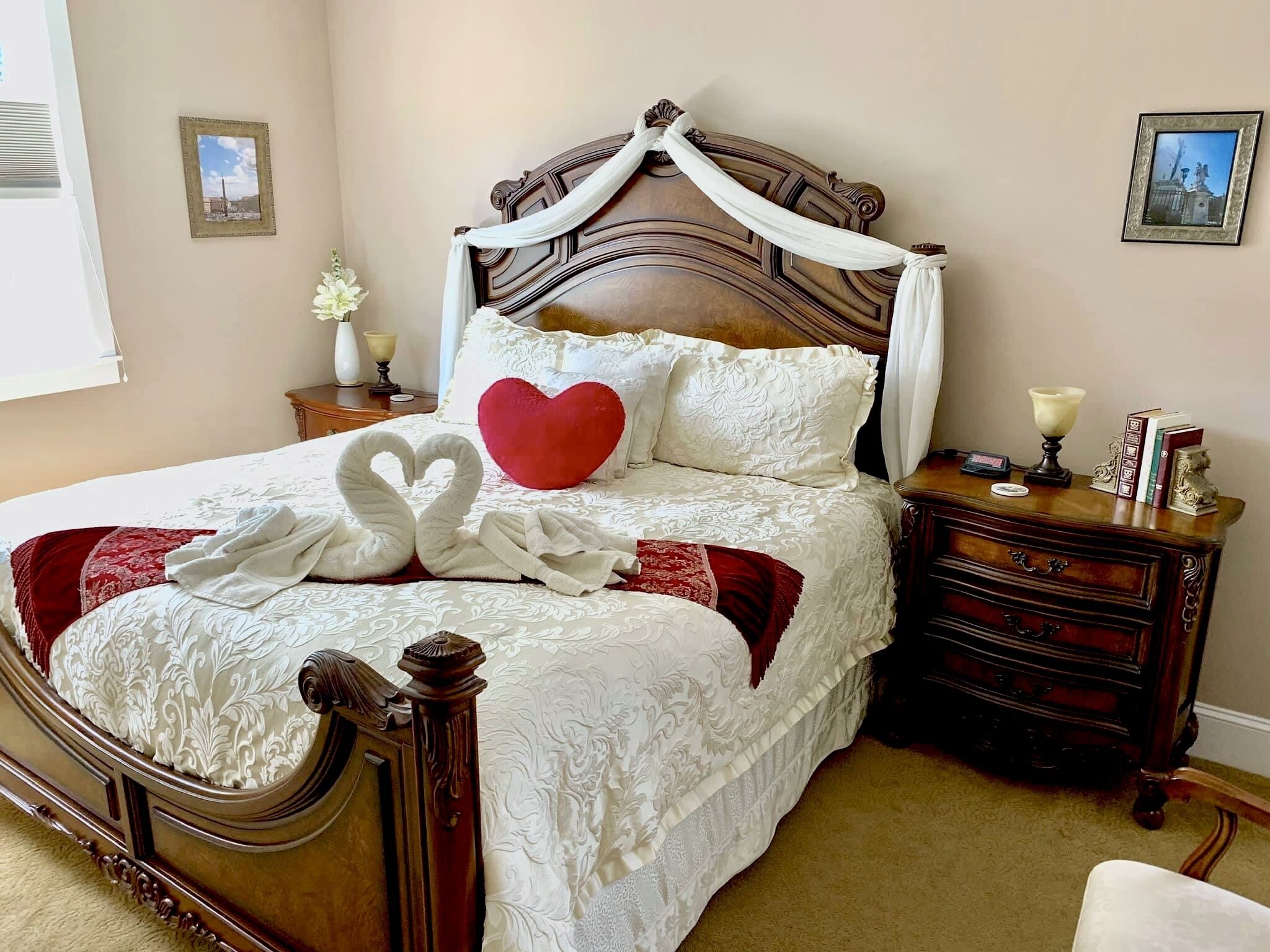 A neatly made bed with a heart-shaped pillow and two swan-shaped towels, a wooden nightstand with a lamp and books, and a wall with two framed pictures.