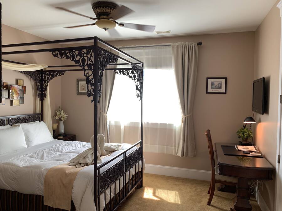 A four-poster bed with a ceiling fan, a desk with a lamp, and a window with curtains.