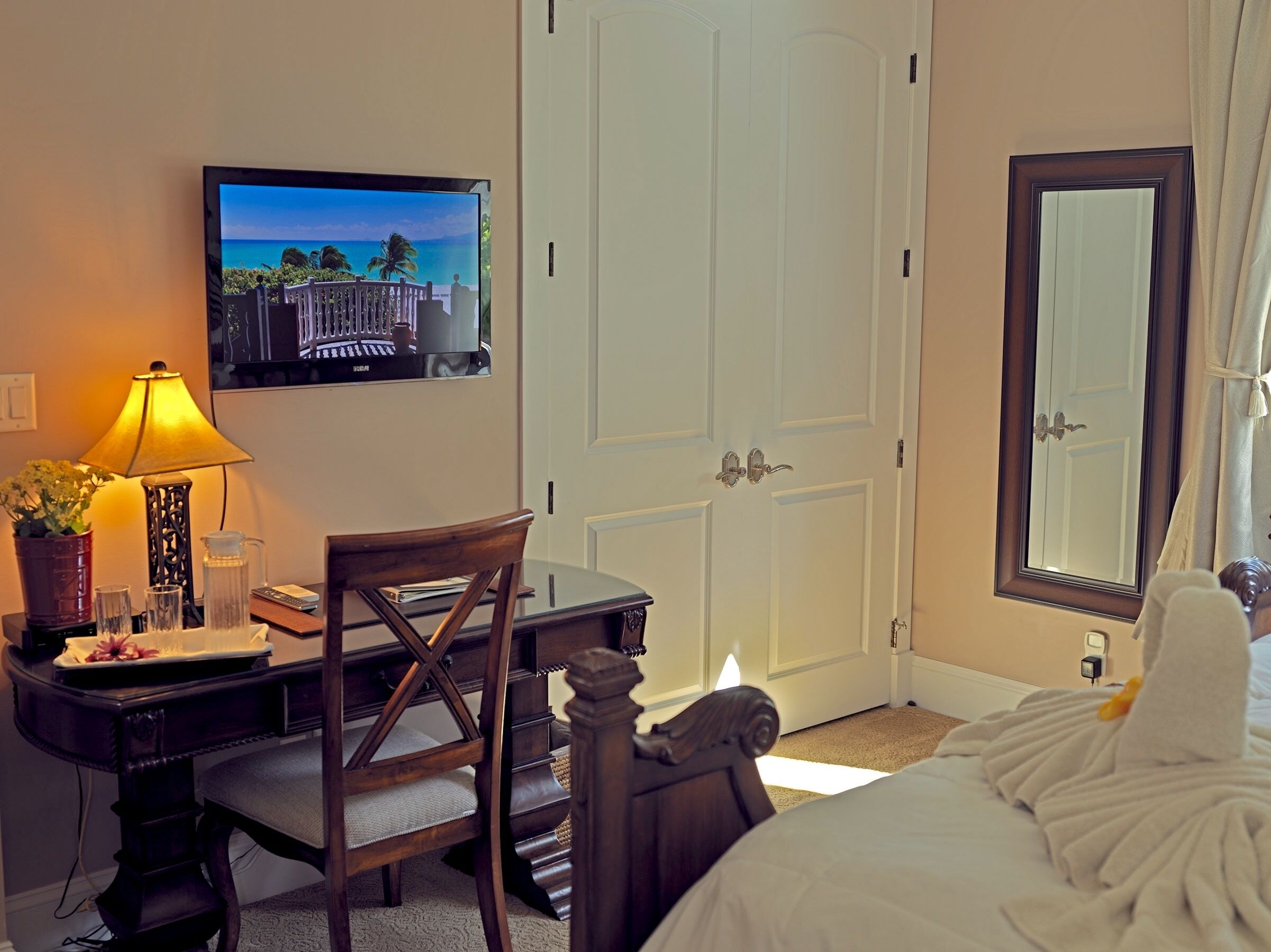 A hotel room with a bed, a desk with a chair, a television mounted on the wall, and a large mirror.