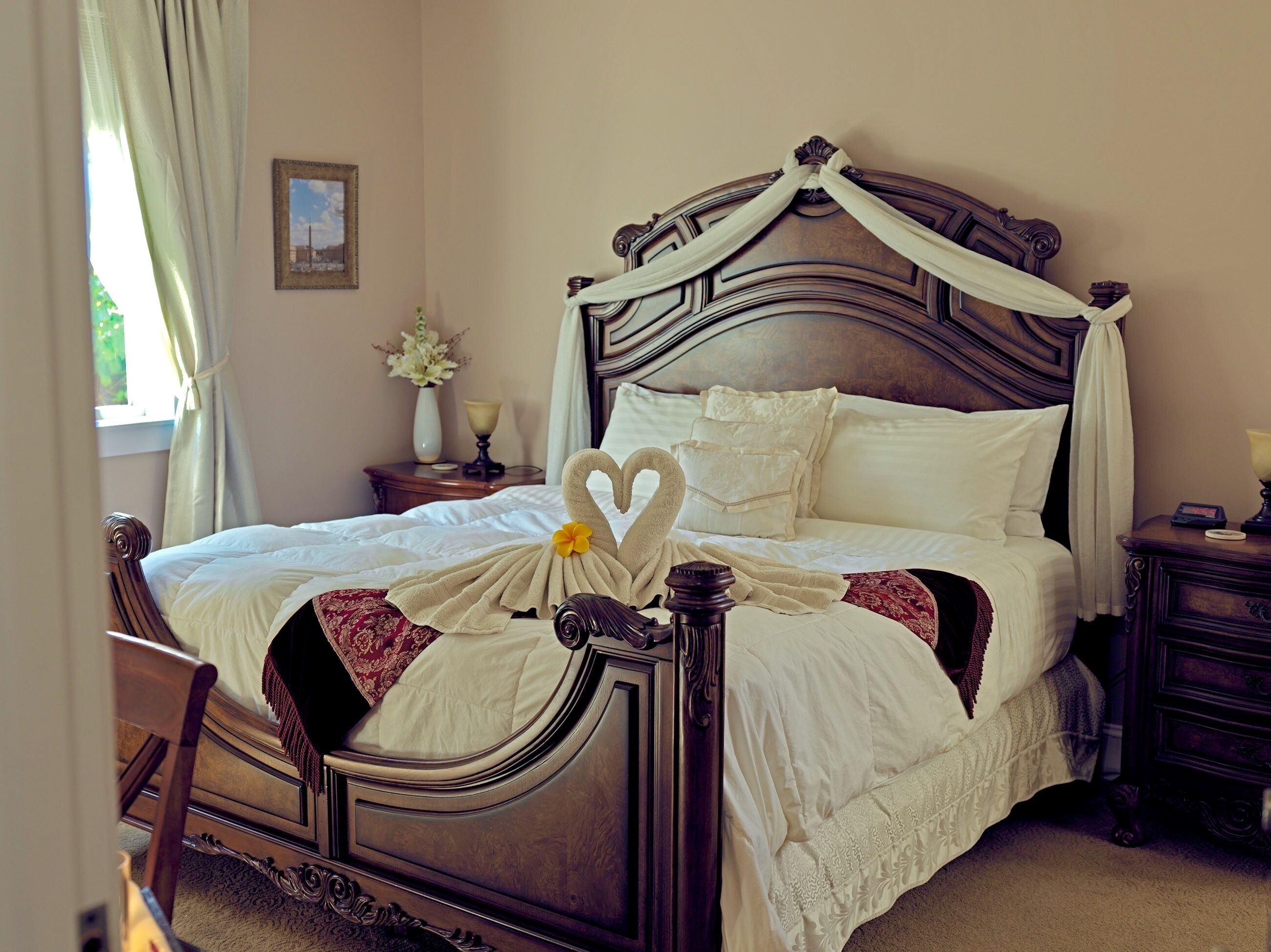 A neatly made bed with a heart-shaped towel arrangement, a wooden headboard, and a bedside table with a lamp and a vase of flowers.