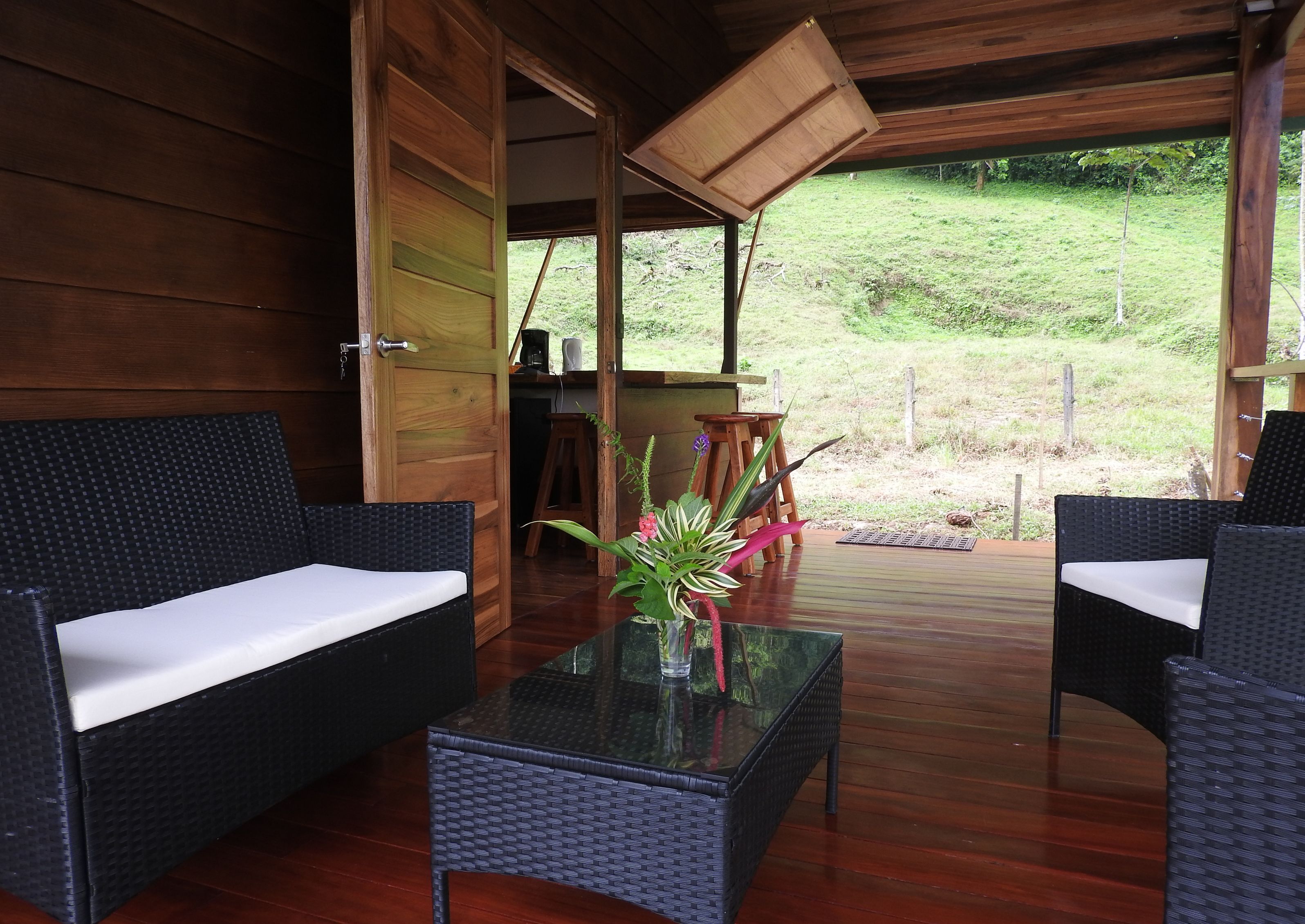 Une cabane en bois avec des meubles en osier, une table en verre et une vue sur un espace verdoyant.