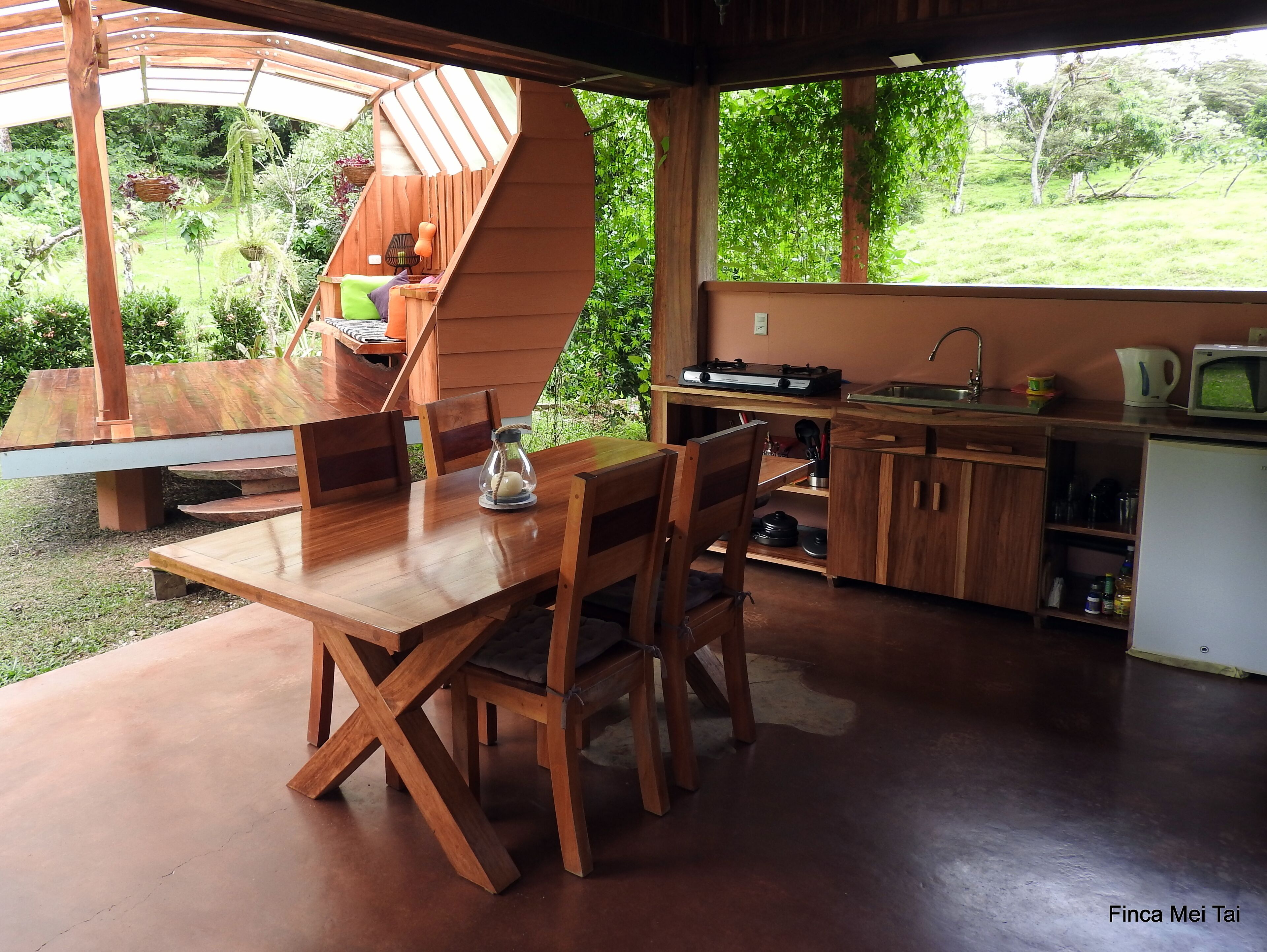 Uma área de jantar rústica em madeira, com uma escada em espiral que leva a um mezanino, uma cozinha com comodidades básicas e vista para um jardim exuberante.