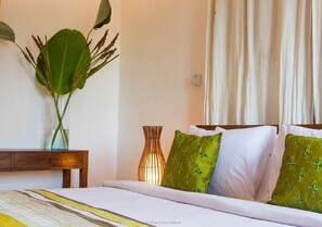 A neatly made bed with white linens and green pillows, a bedside table with a vase of plants, and a wall-mounted light fixture.