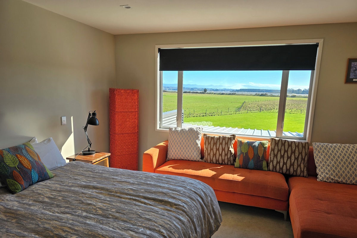 A bedroom with a bed, a sofa, and a large window offering a view of a green field.