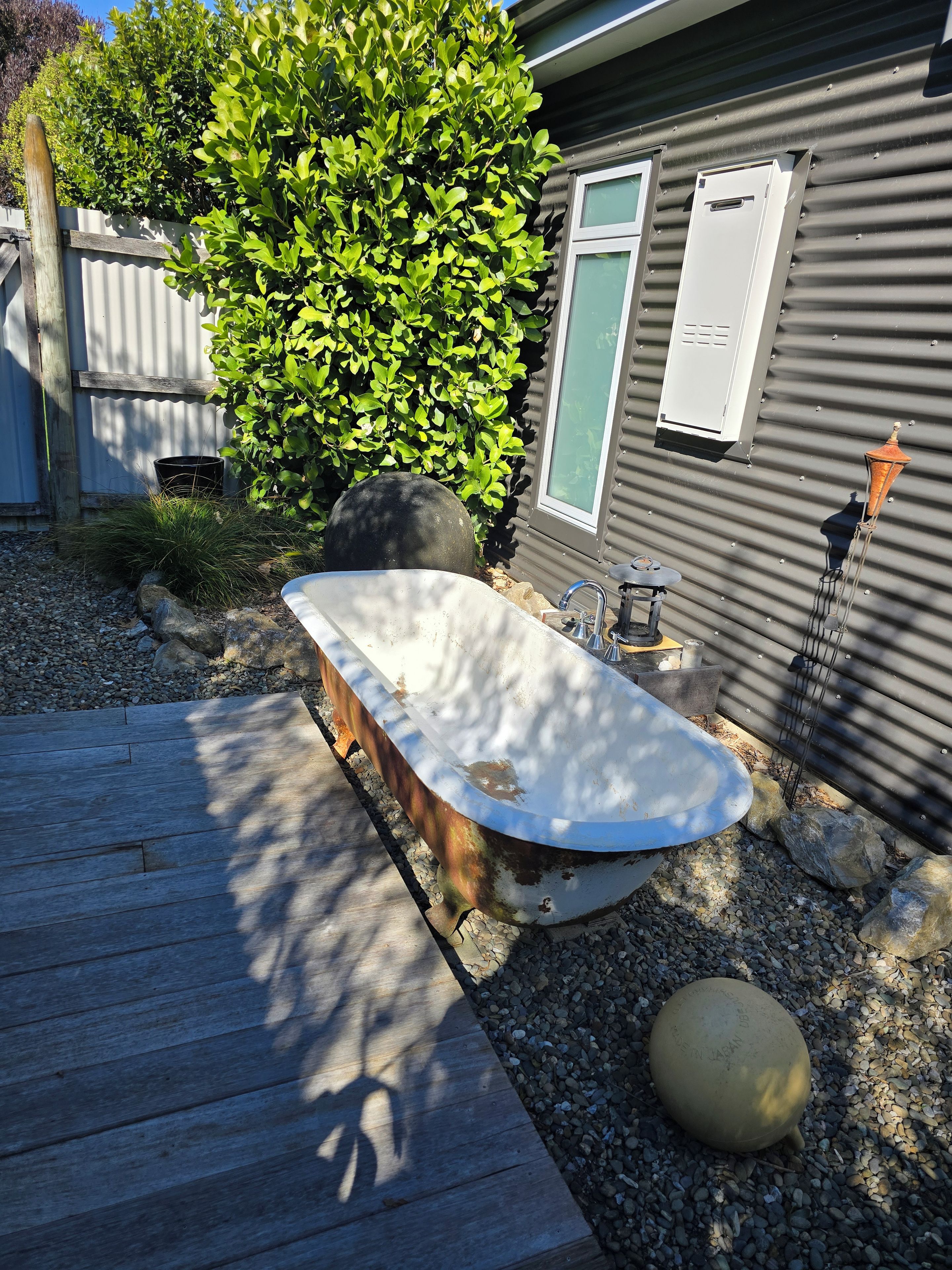 A wooden deck with a large, rustic bathtub, a small garden area with rocks and a potted plant, and a metal fence.